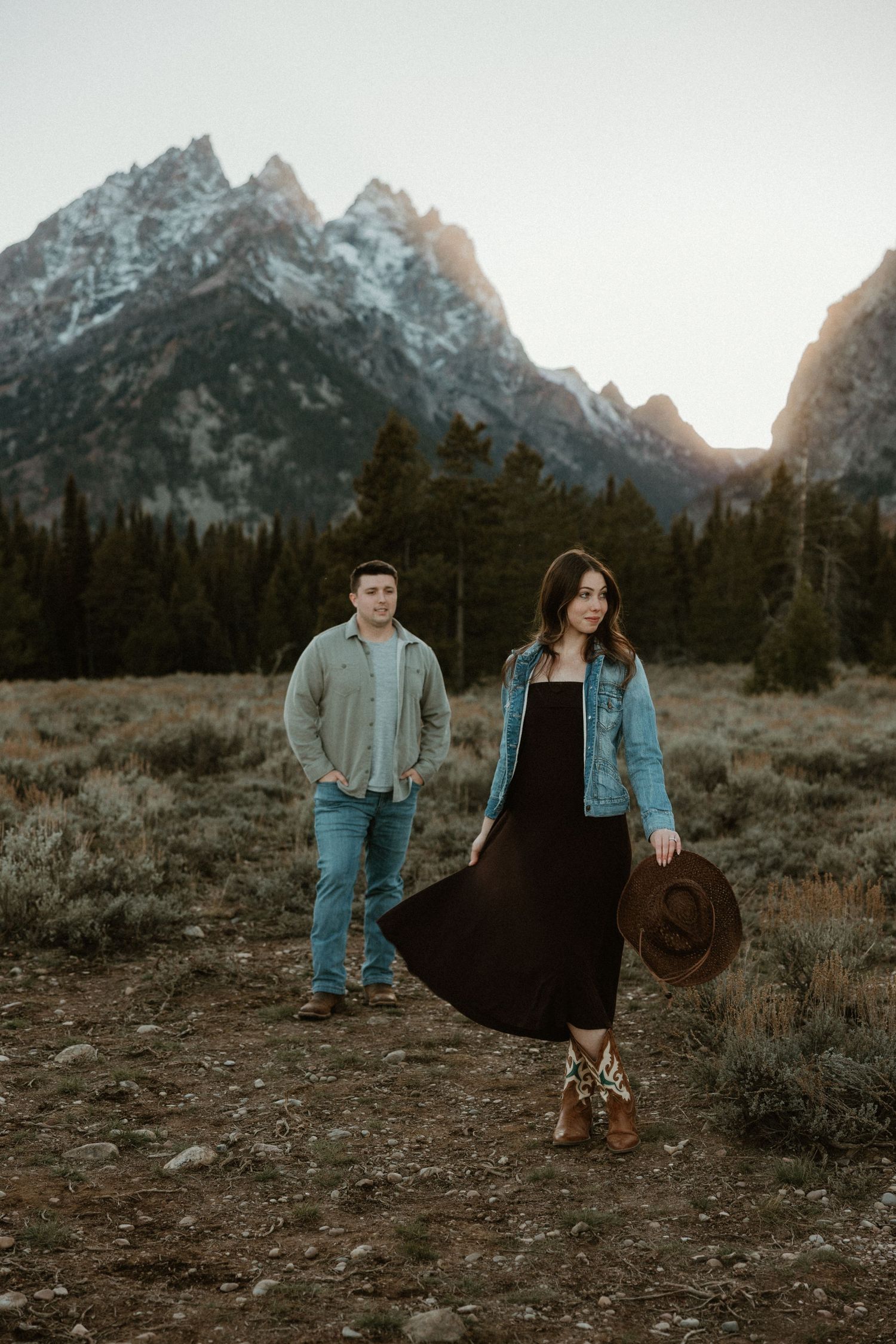 Couple poses in casual western wear against majestic mountain backdrop during sunset in Grand Teton National Park.