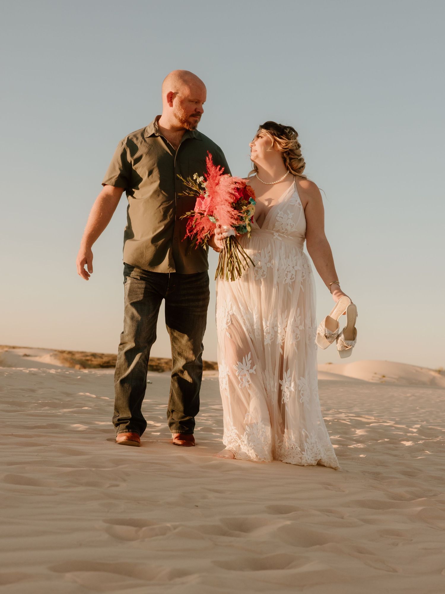 A couple walks hand in hand on desert sand dunes with a bright red floral bouquet.