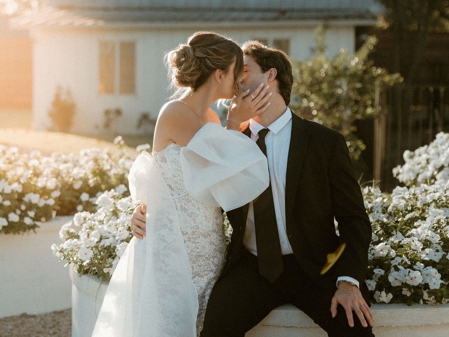 Elegant wedding couple embraces during golden hour with white flowers in the background at an outdoor venue.