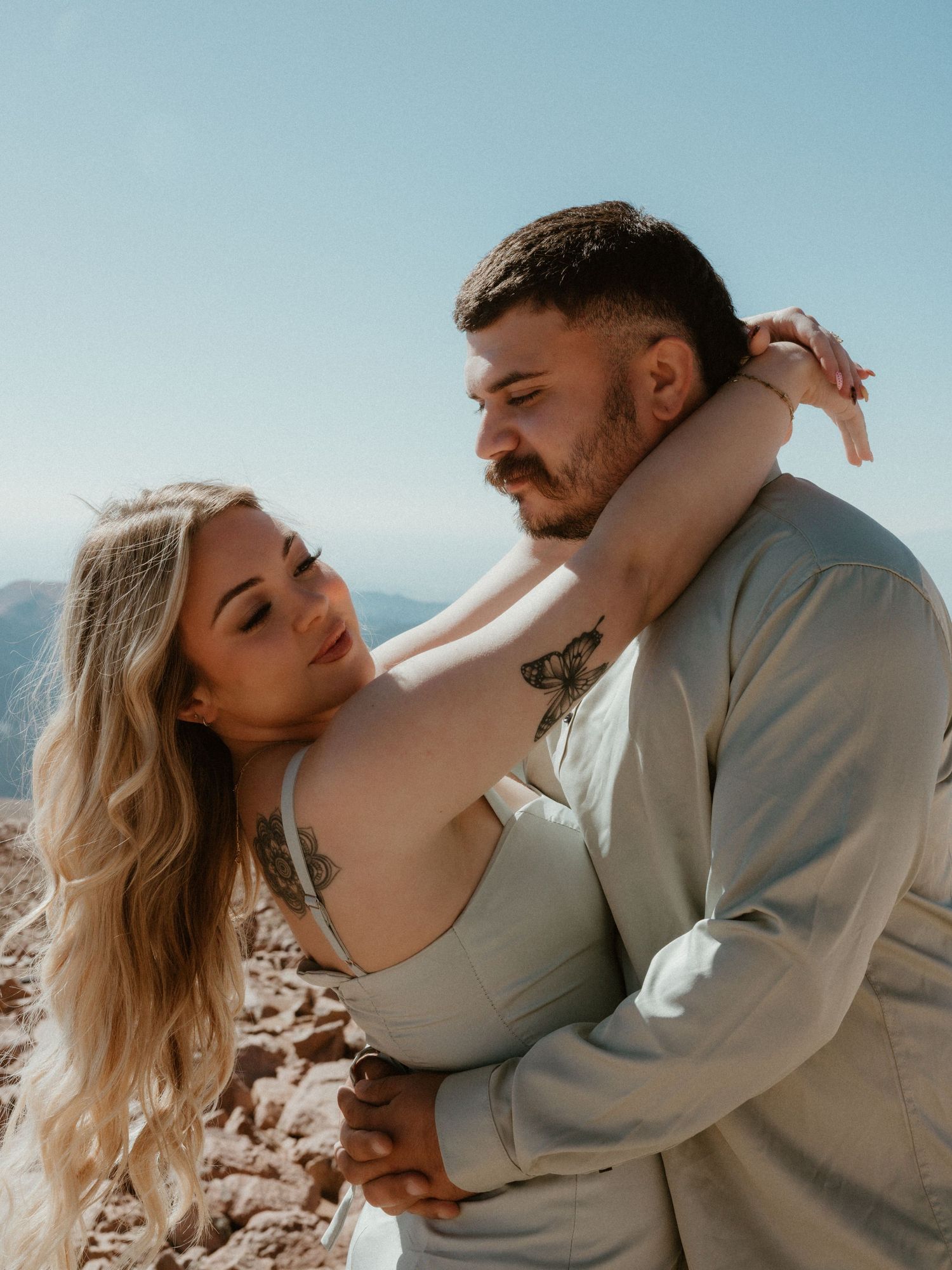 A romantic couple embraces on a mountain summit during a sunny day with scenic views in the background.