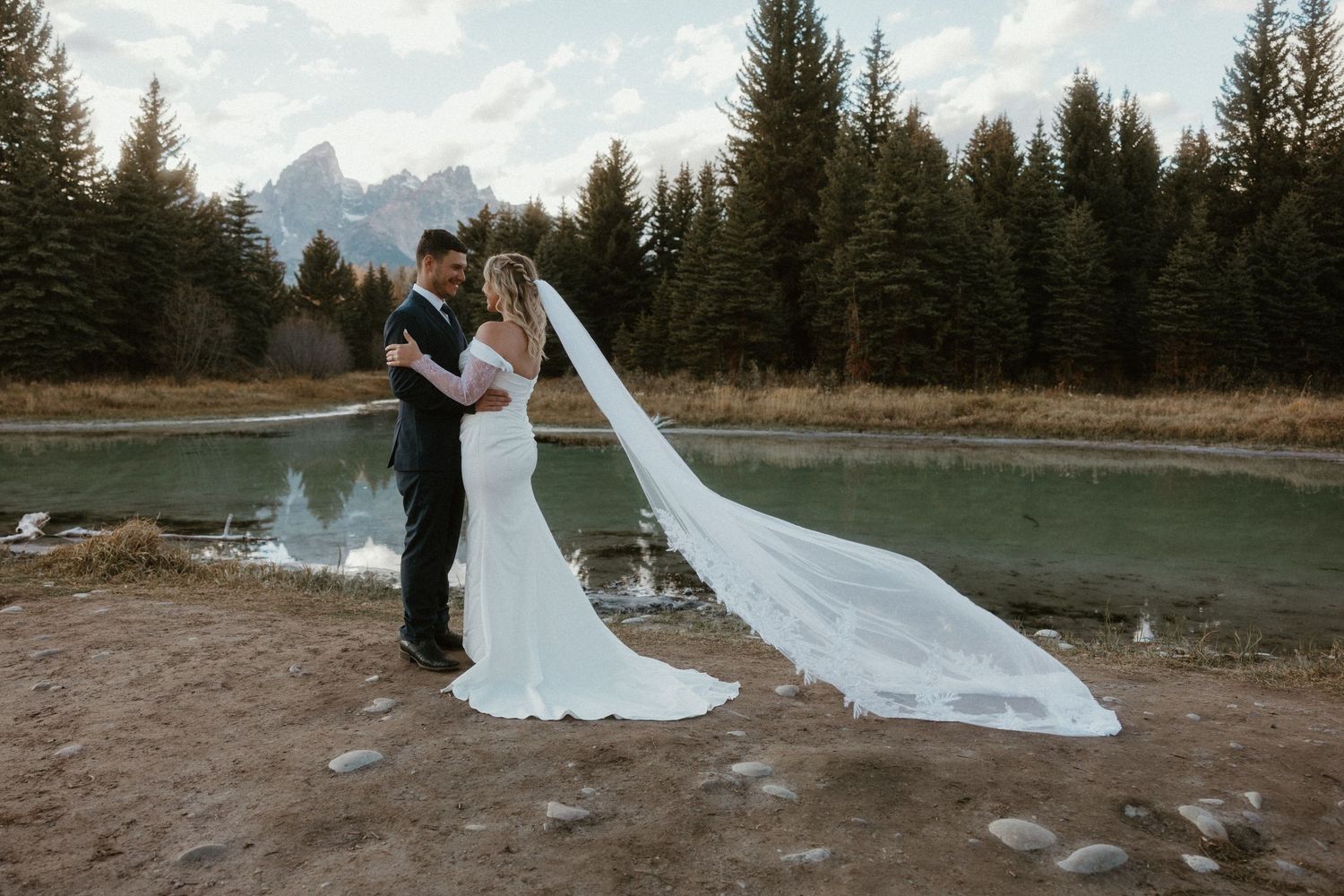 A wedding couple poses against a scenic mountain backdrop with evergreen trees and a flowing white dress train.