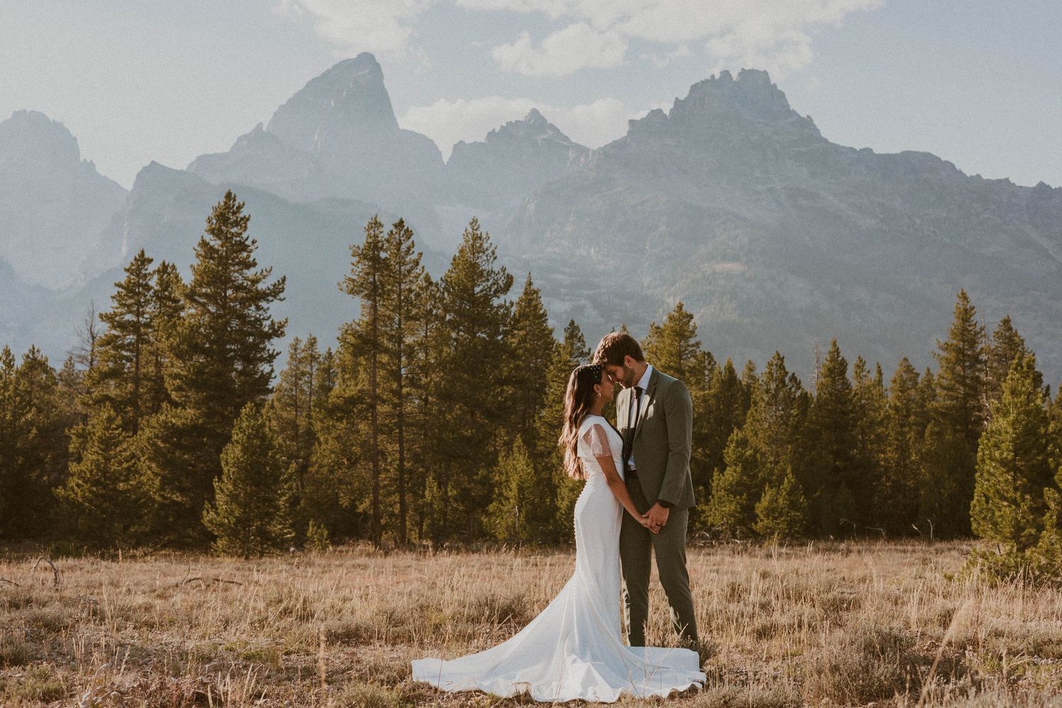 A couple shares an intimate moment in a mountain meadow with dramatic peaks in the background.