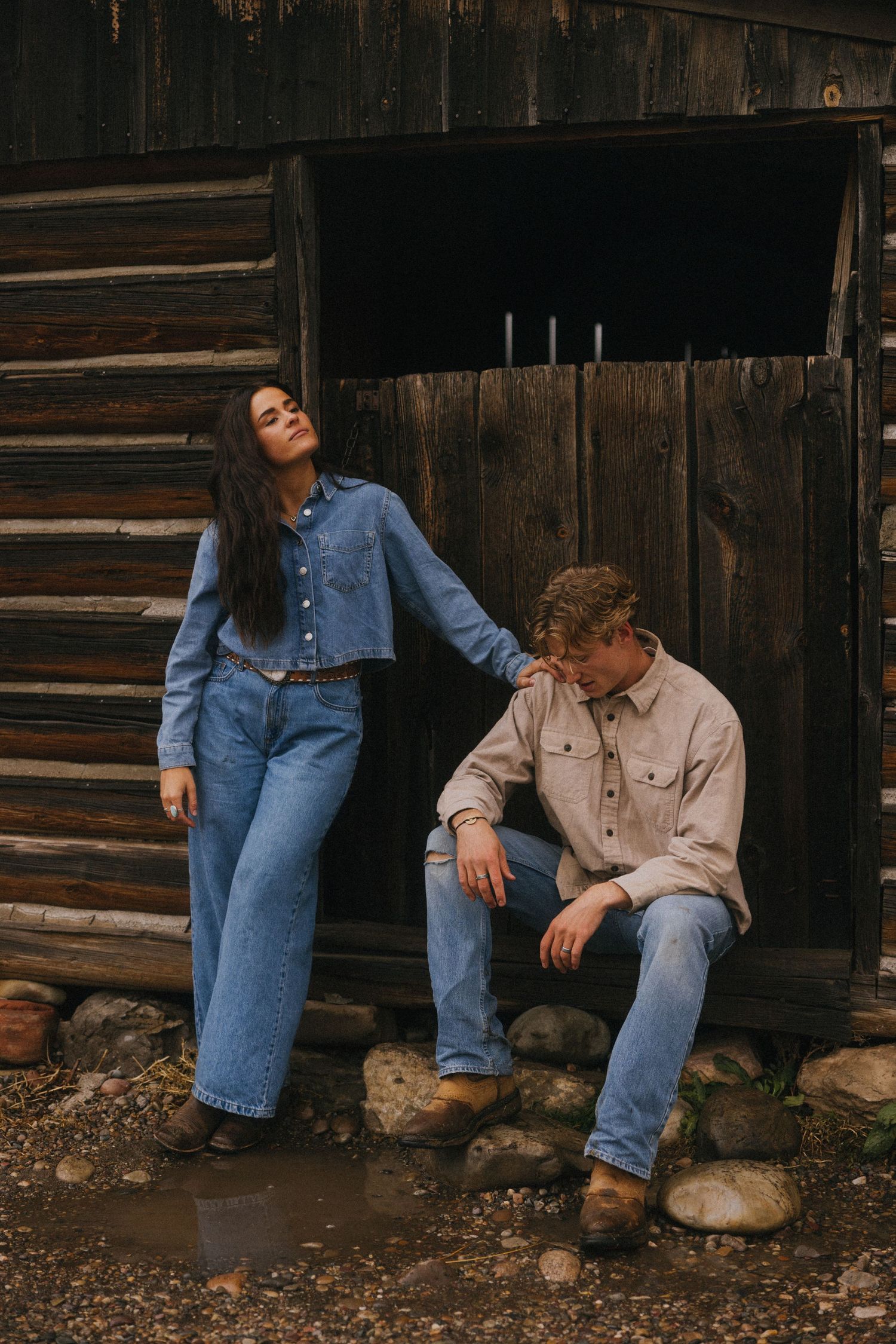 A couple sits casually against a rustic wooden wall in matching denim outfits.