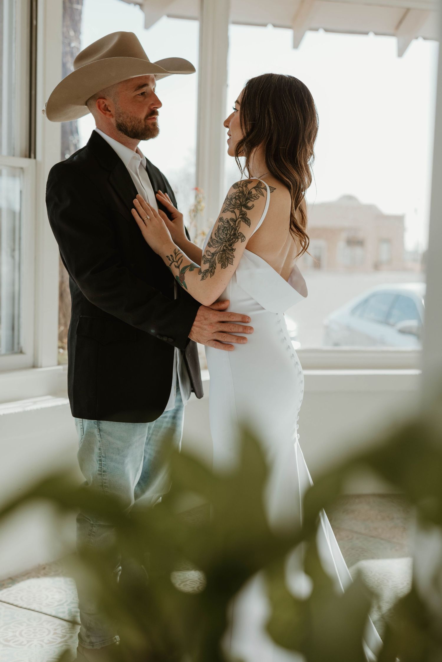 Romantic wedding couple embracing near bright windows with indoor greenery.