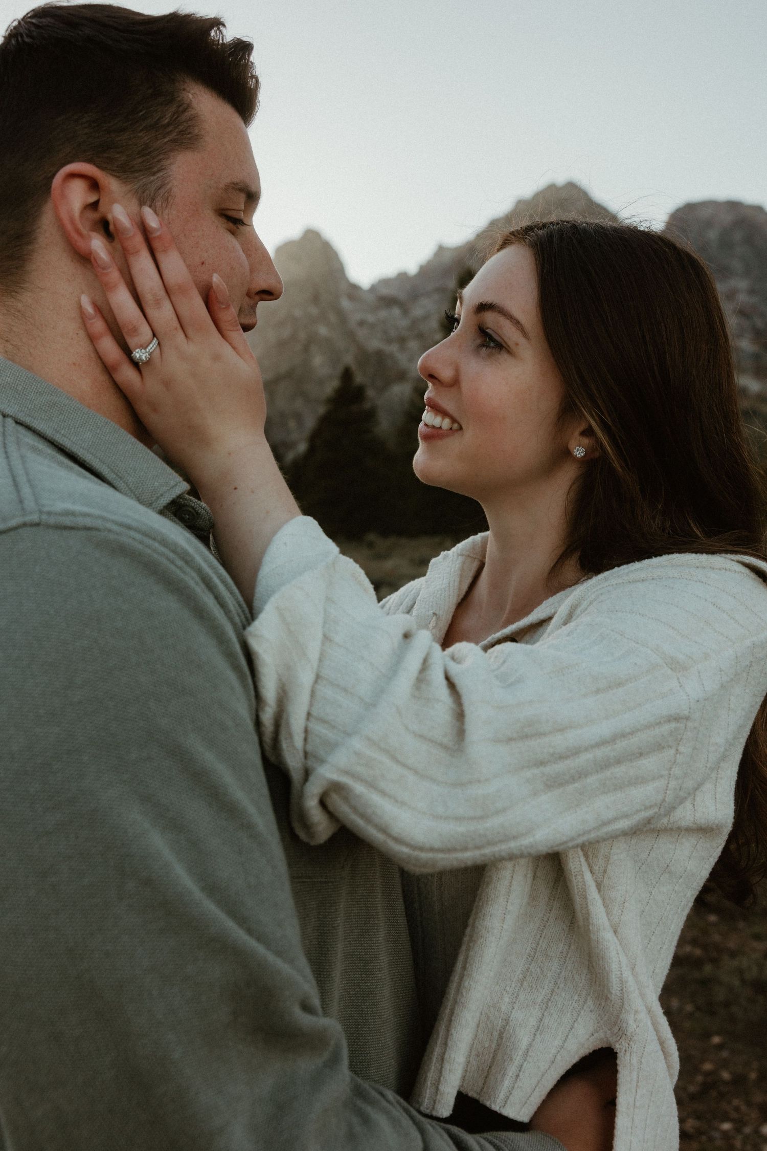 Tender moment between couple in white sweater and grey jacket with mountains in background.