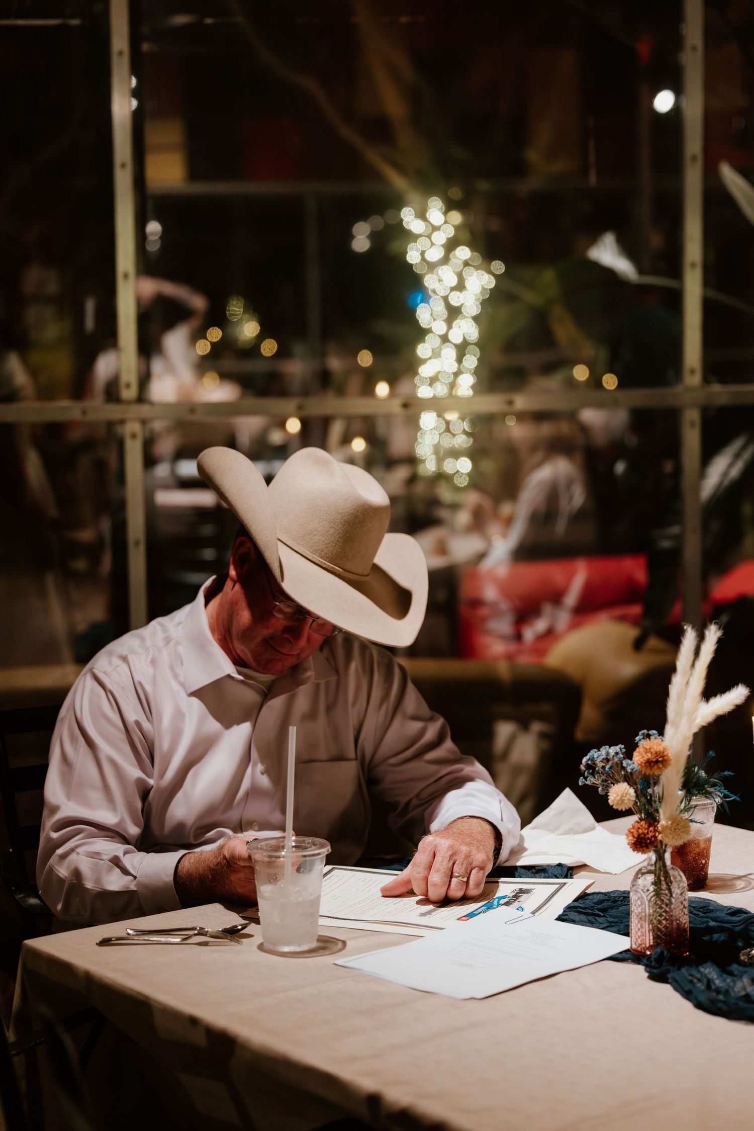 A guest in a cowboy hat signs the guestbook at a wedding reception with twinkling lights in the background.