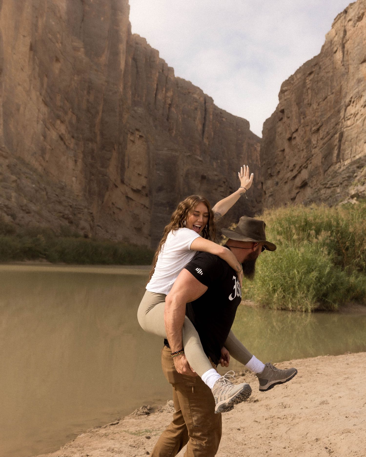 Two people playfully interact near a scenic canyon wall beside a river.