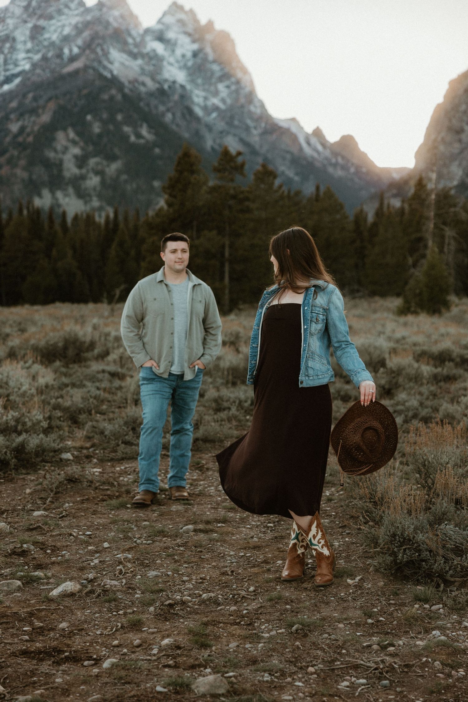 Couple in casual attire standing apart in mountain field during sunset.