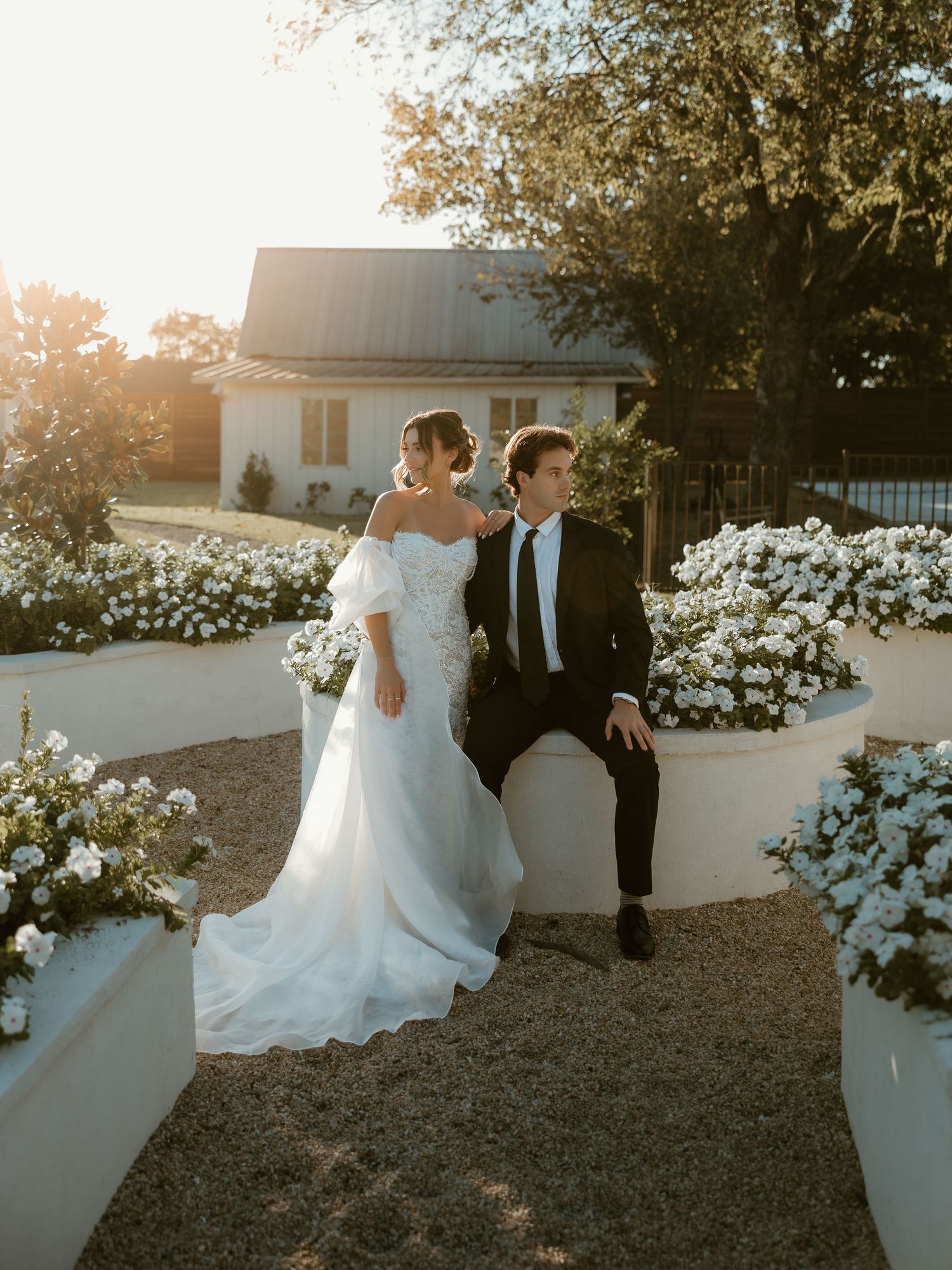Couple in wedding attire walking together through a garden path with white flowers at sunset.