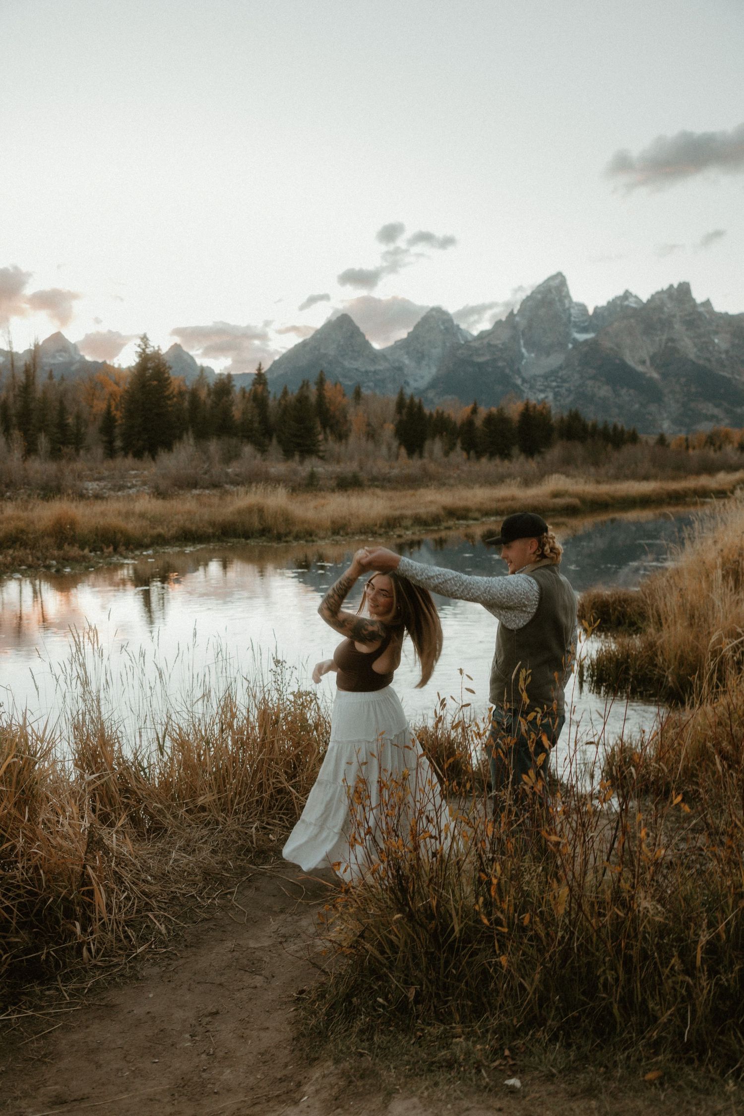 Playful couple dancing by a mountain lake surrounded by autumn grasses and snow-capped peaks.