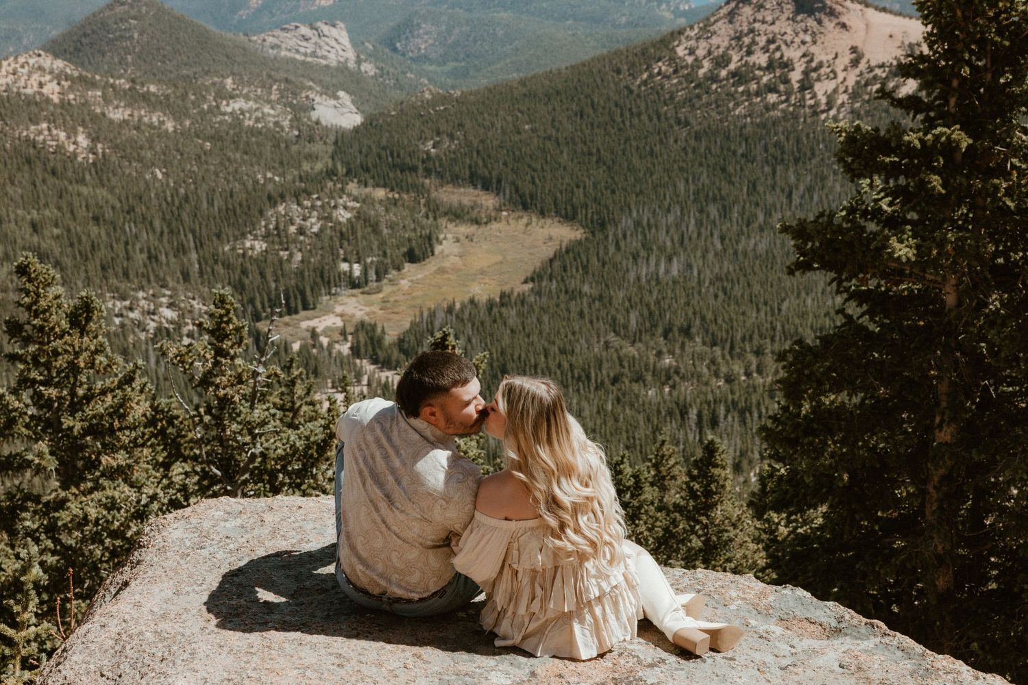 Couple sits together on rocky mountain ledge overlooking vast forest valley below.