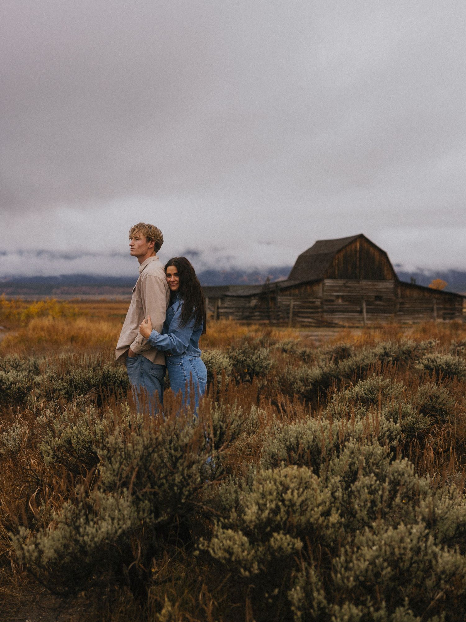 A couple embraces in a field with mountains and an old barn in the distance.