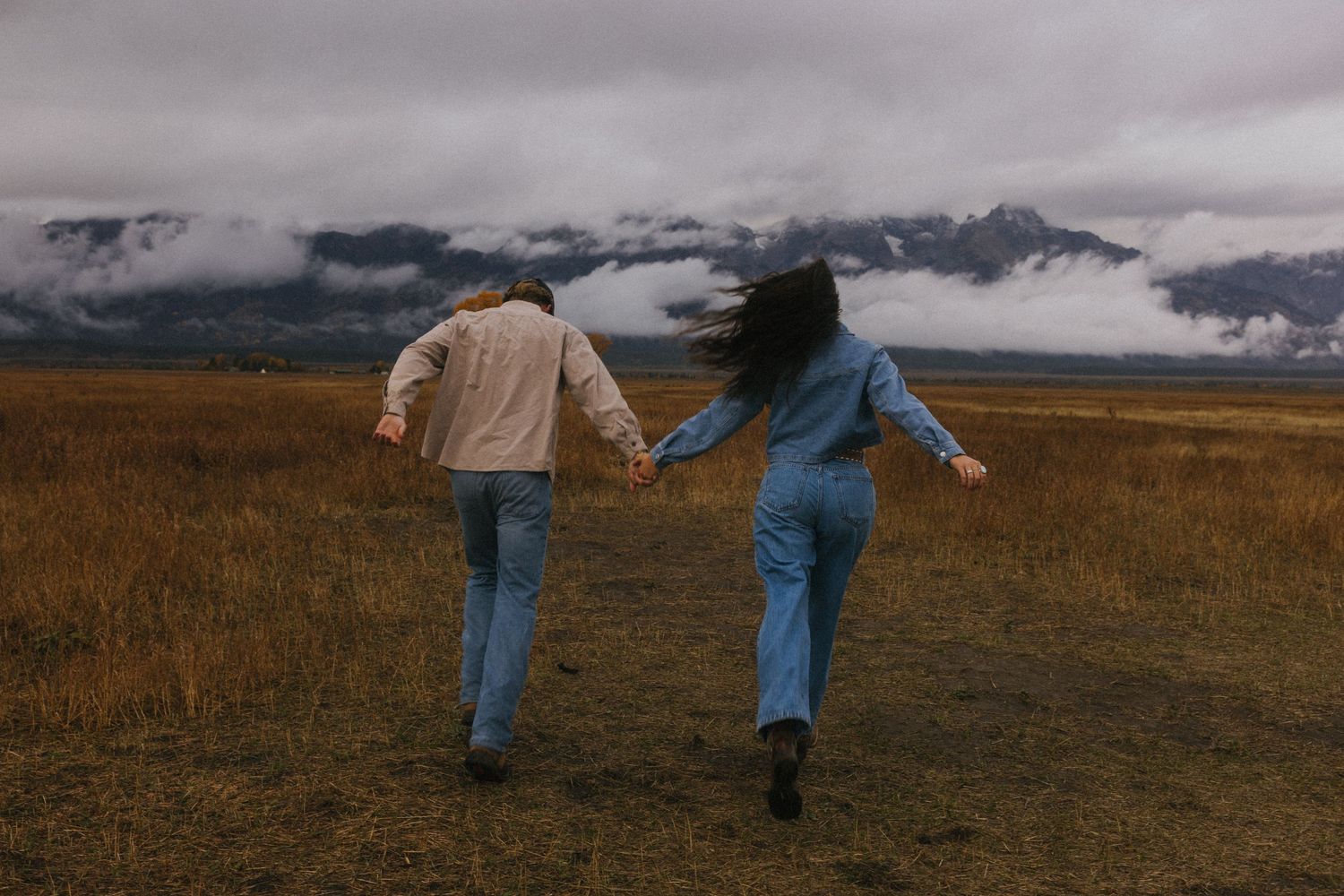 Two people hold hands while running through autumn field with cloudy mountains in background.