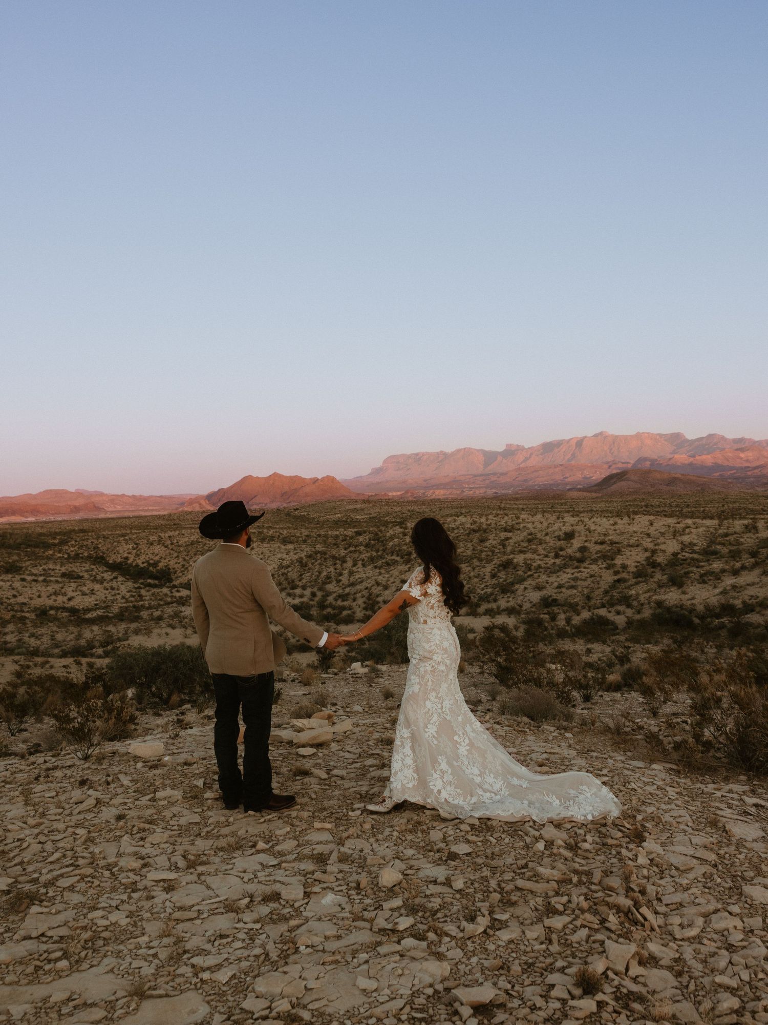 A couple in wedding attire poses against a desert landscape with mountains at sunset.