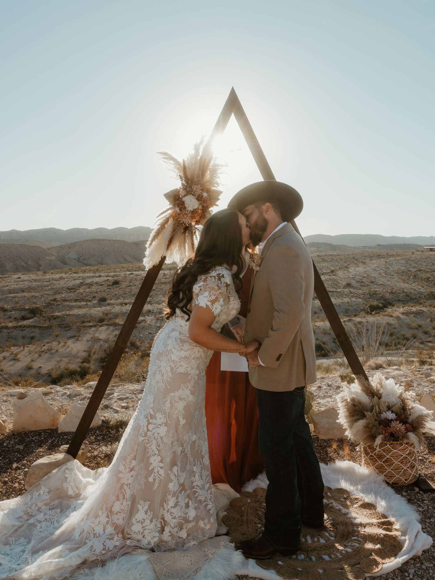 A desert wedding ceremony setup with wooden arch and flowing white dress against rocky terrain.