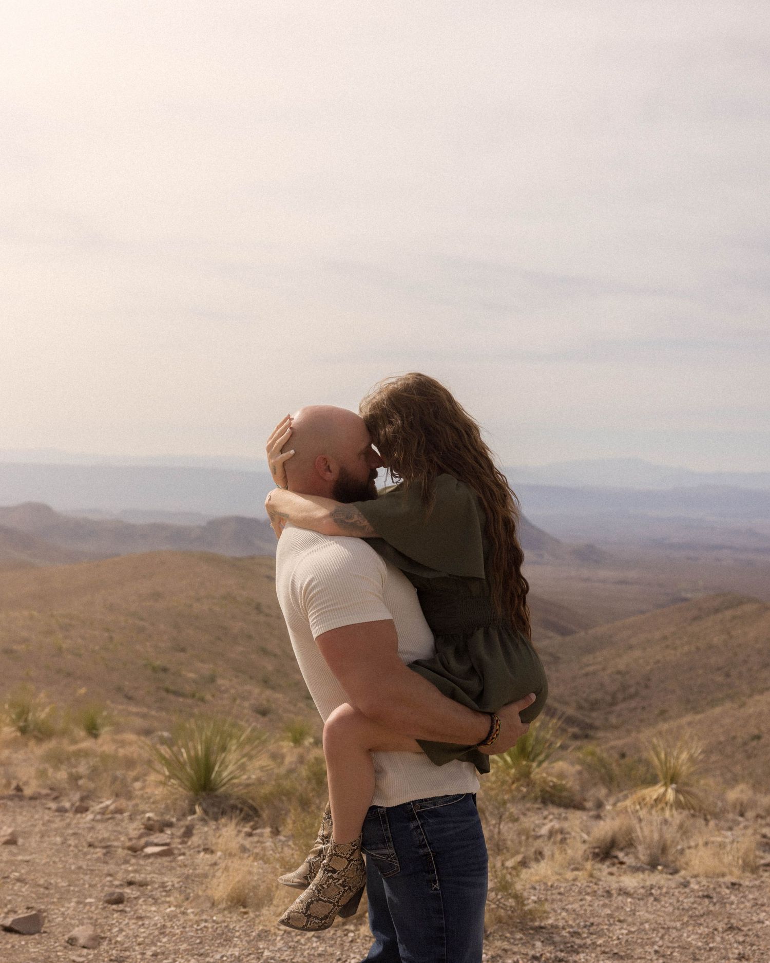 A couple embraces while overlooking a vast desert mountain landscape during sunset.