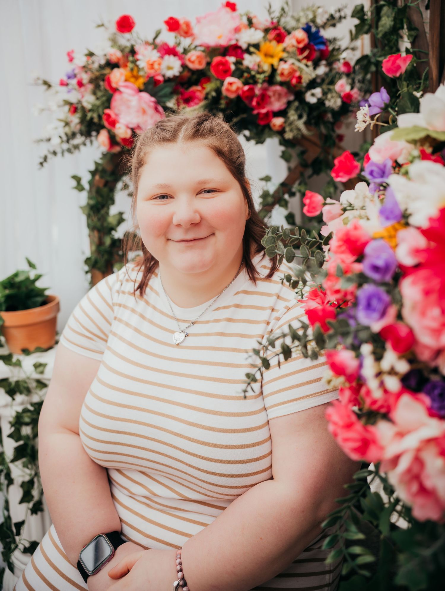 A person in a striped shirt sits surrounded by vibrant pink and purple flowers in an outdoor garden setting.