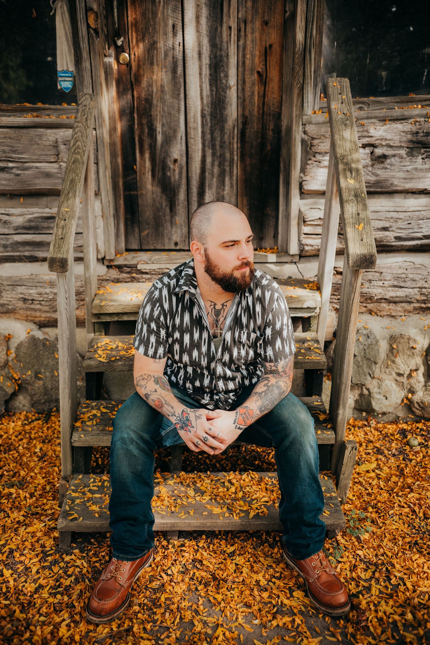 Person with tattoos and beard sits on wooden steps surrounded by fallen autumn leaves in rustic setting.