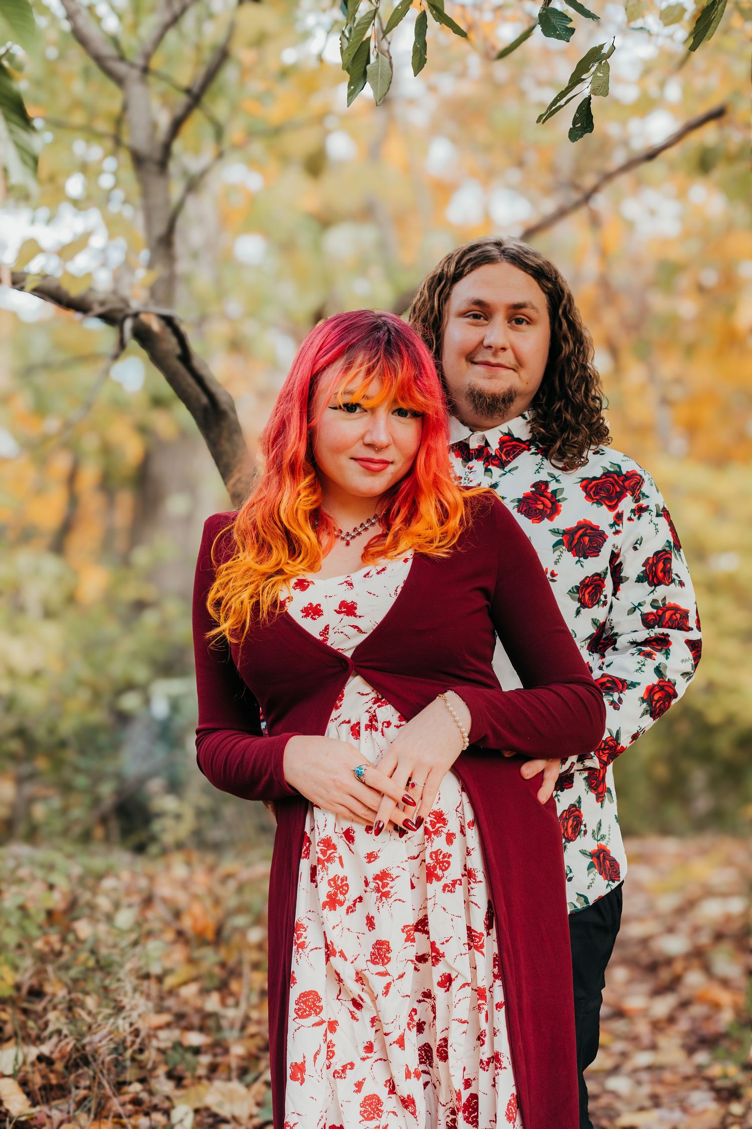 A couple in coordinating burgundy and floral outfits poses together during an autumn photoshoot with golden leaves.