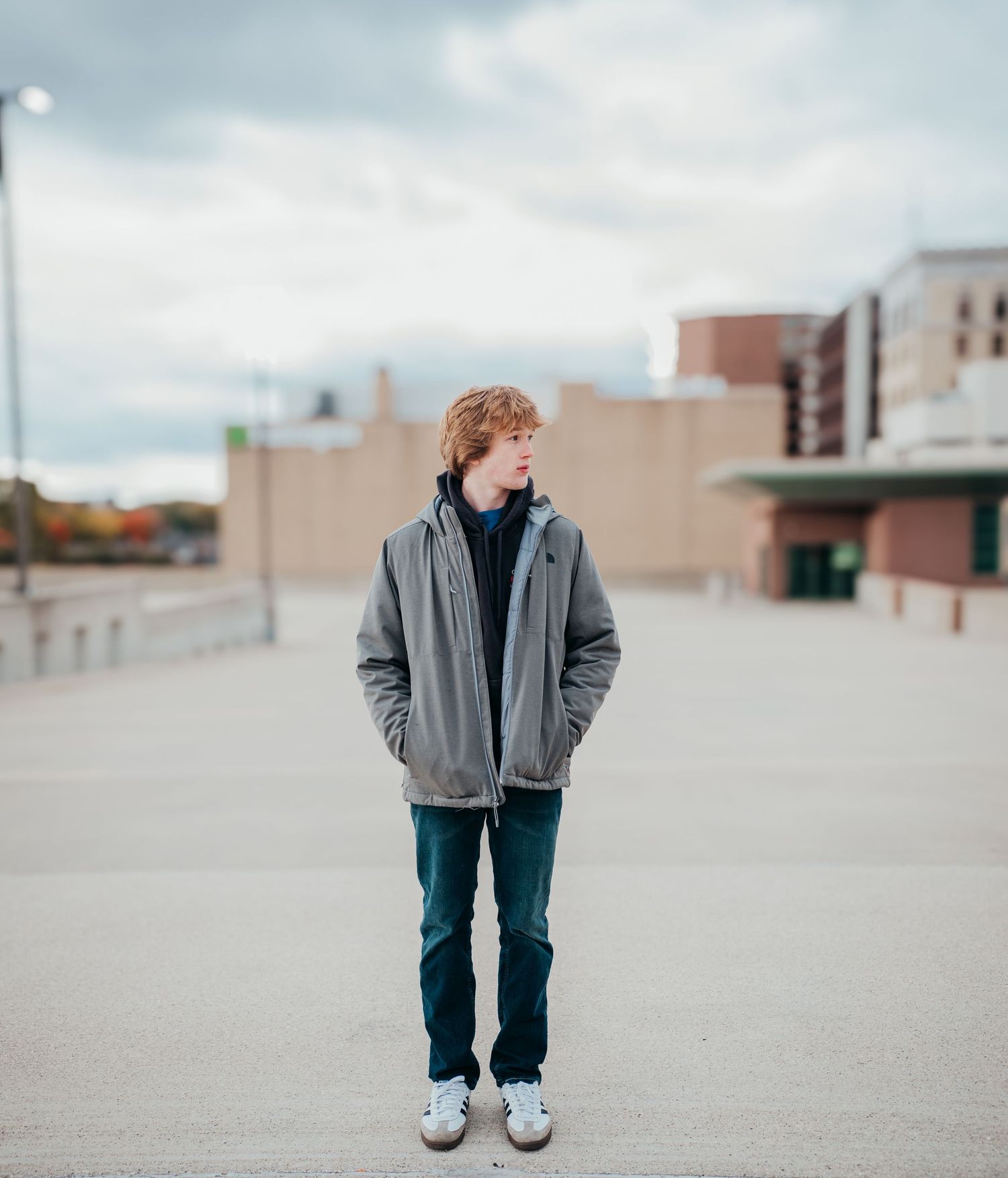 A person in a gray jacket and jeans stands alone on an urban rooftop area with buildings in the background.