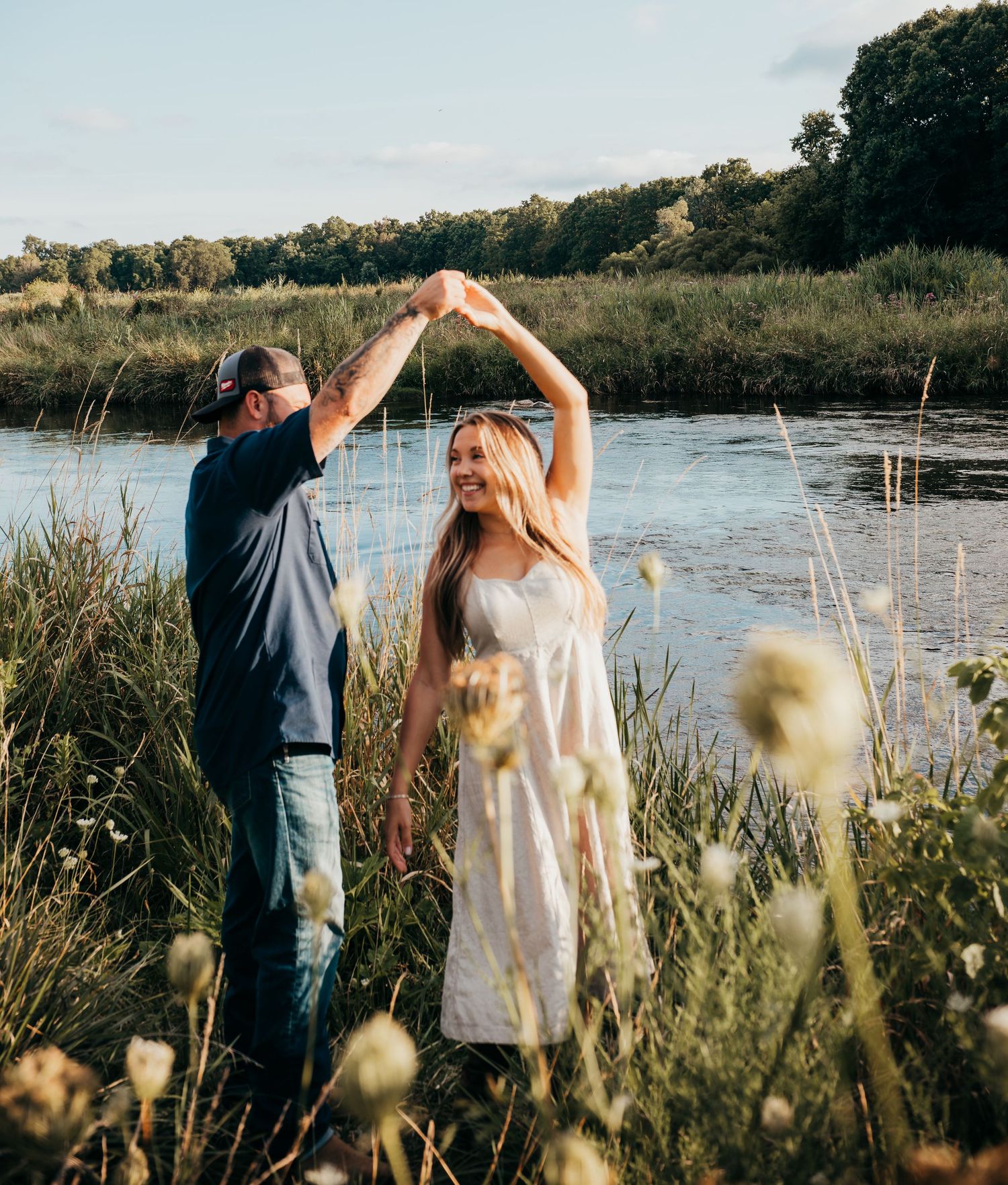 A couple dances together in a romantic outdoor setting by a lake at sunset surrounded by tall grass and wildflowers.