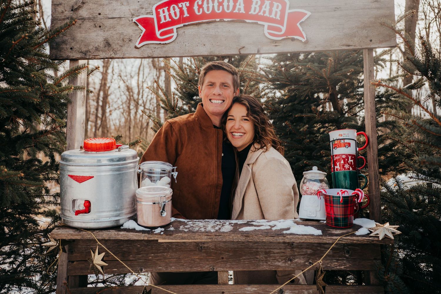 A couple stands behind a rustic hot cocoa bar setup with festive winter decorations and pine trees in the background.