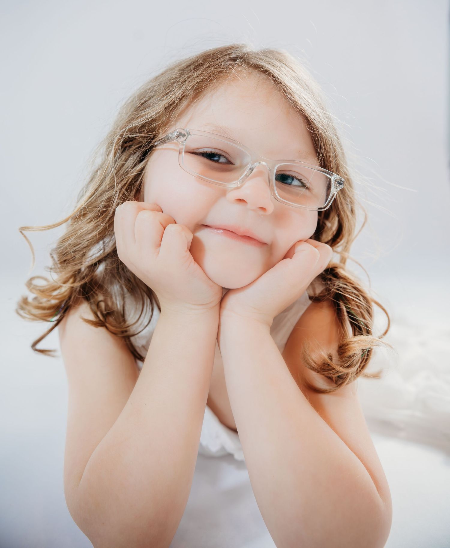 A young child with curly hair and glasses rests their head on their hands while smiling at the camera.