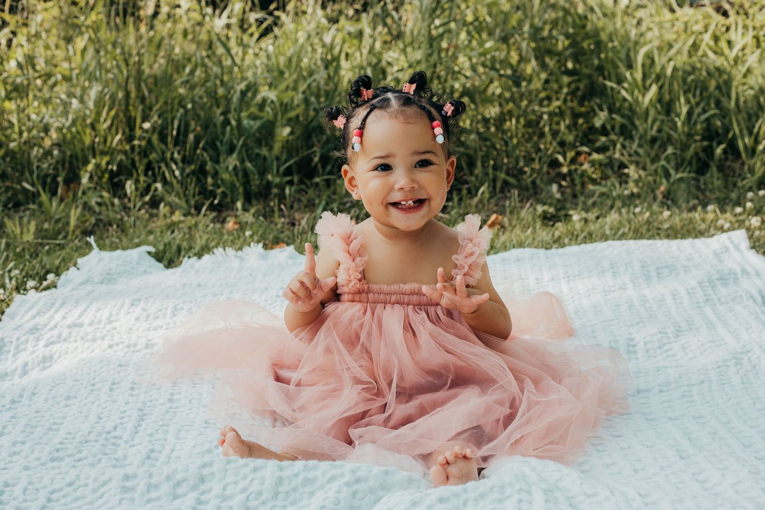 Smiling baby wearing pink tulle dress sits on white blanket outdoors with green foliage in background.