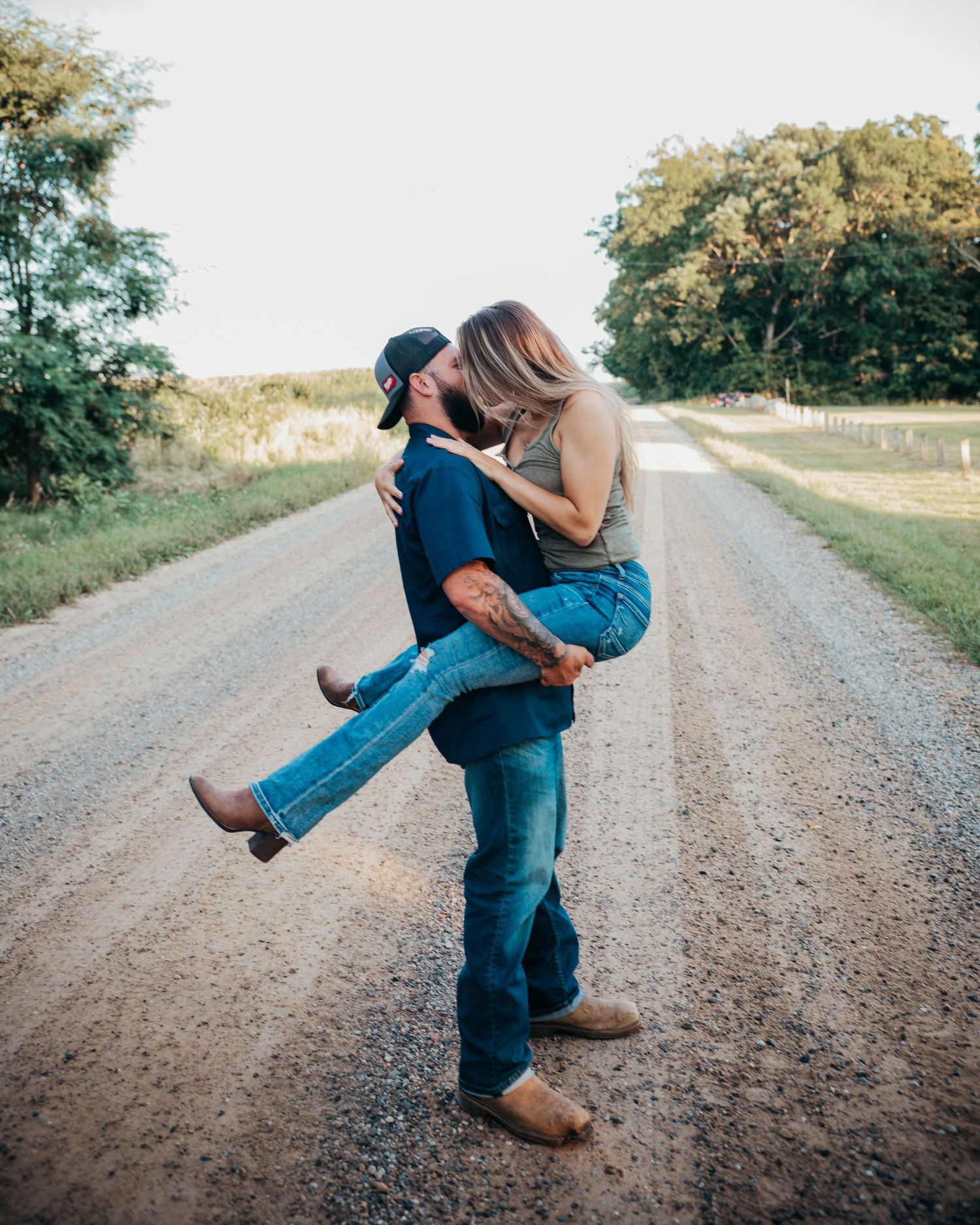 A couple embraces romantically on a country dirt road surrounded by green fields and trees at sunset.