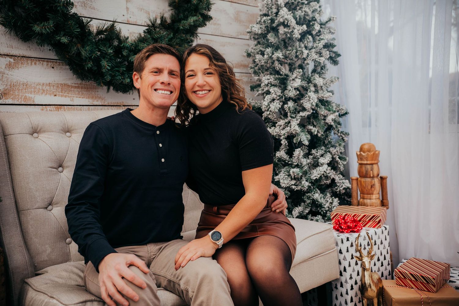 Couple poses together in front of decorated Christmas trees and wrapped presents in a festive holiday setting.