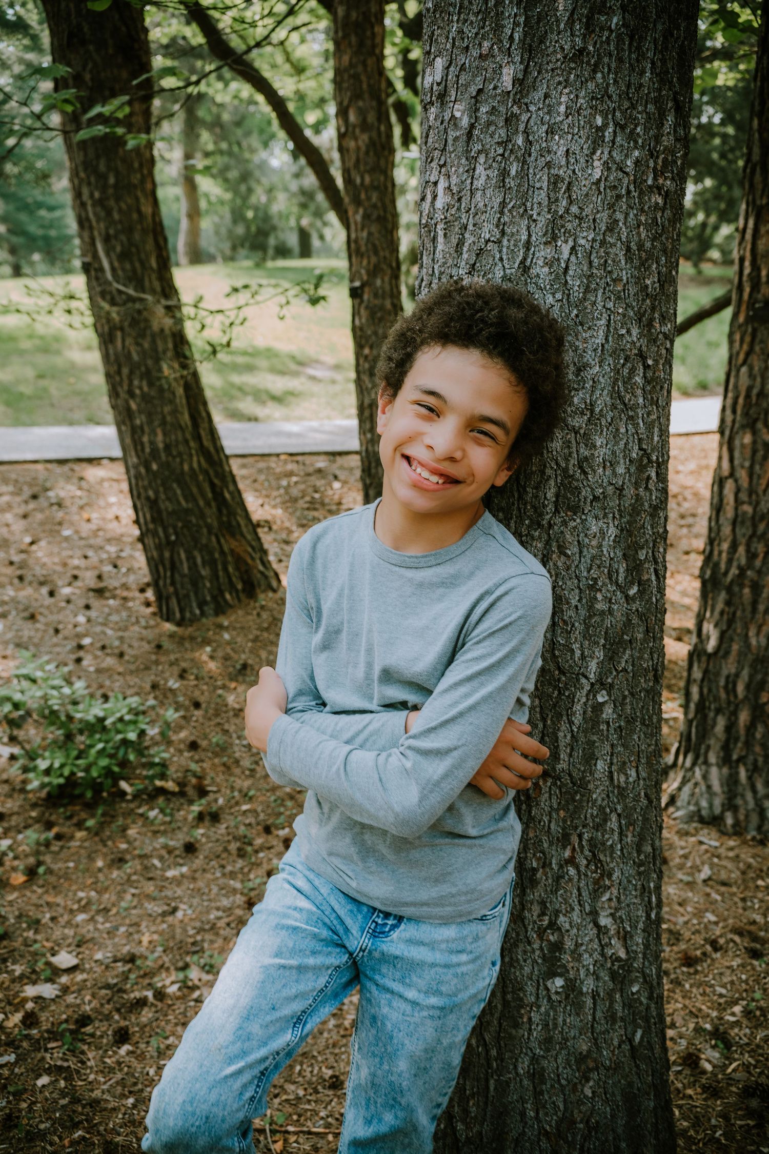Young person in a gray sweater and jeans leans against a tree trunk in a wooded outdoor setting.