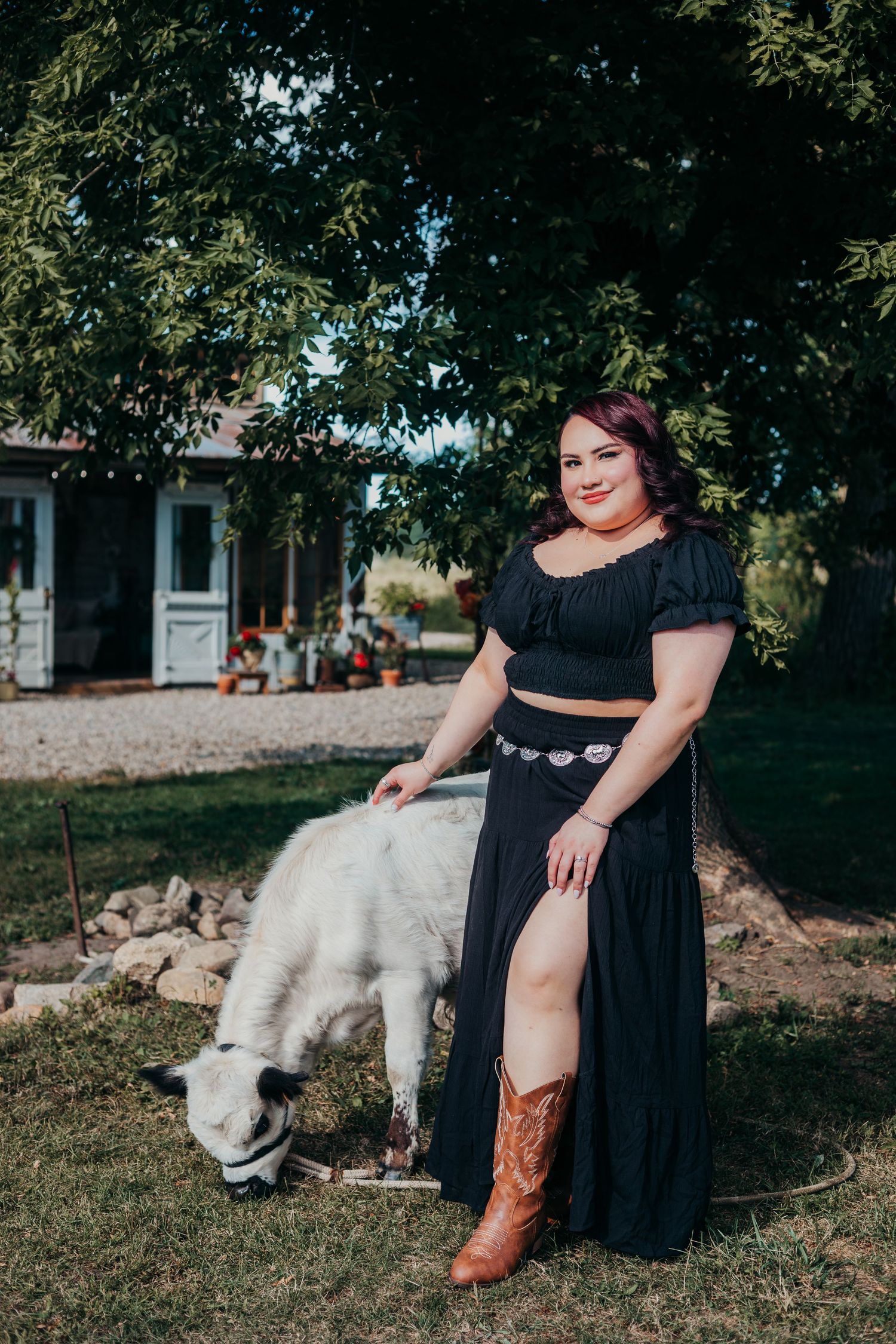 A woman in a black dress and brown cowboy boots poses outdoors near a resting white dog in a garden setting.