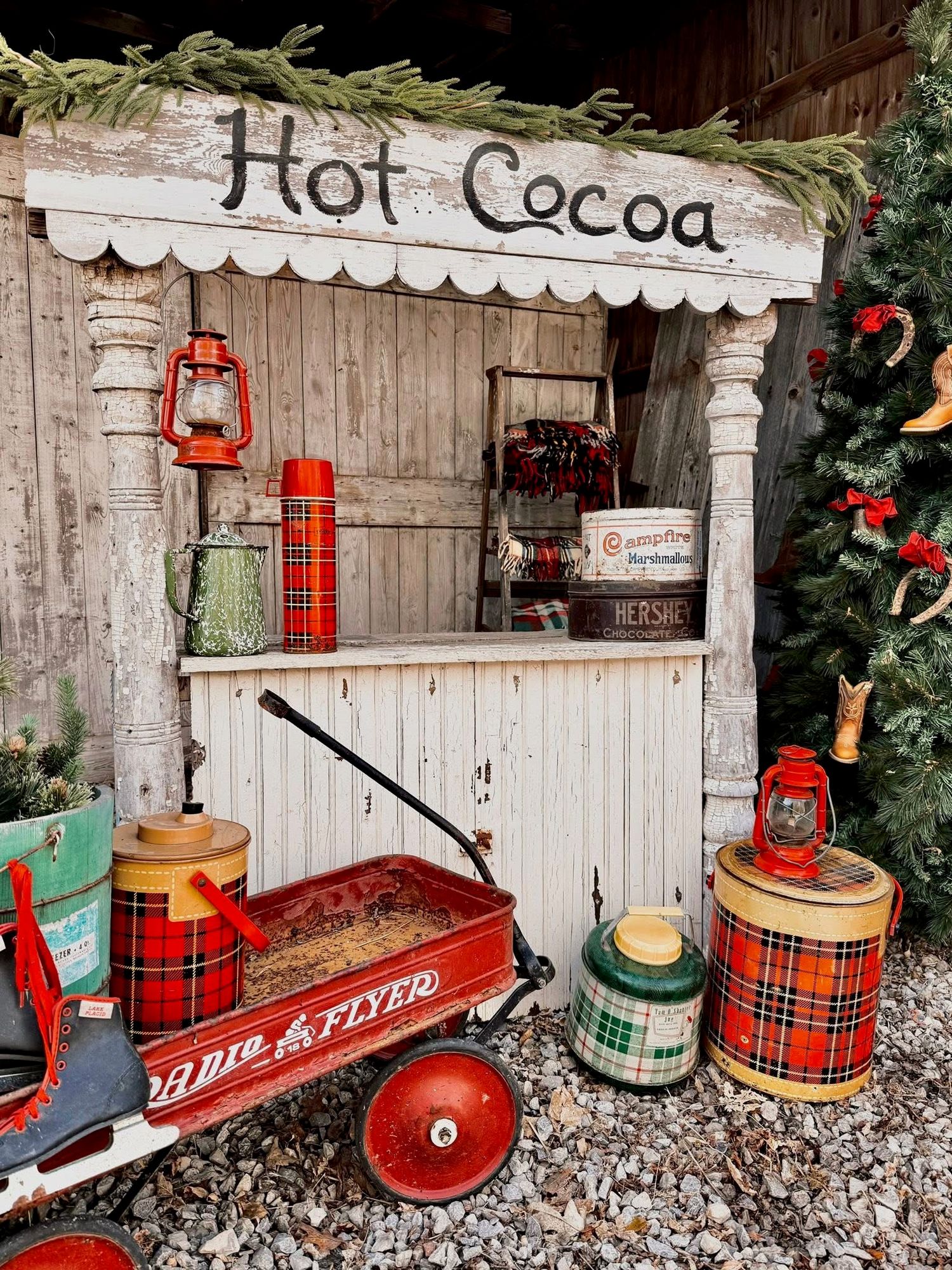 Rustic hot cocoa stand with red lanterns, plaid thermoses, and vintage red wagon decorated for Christmas.