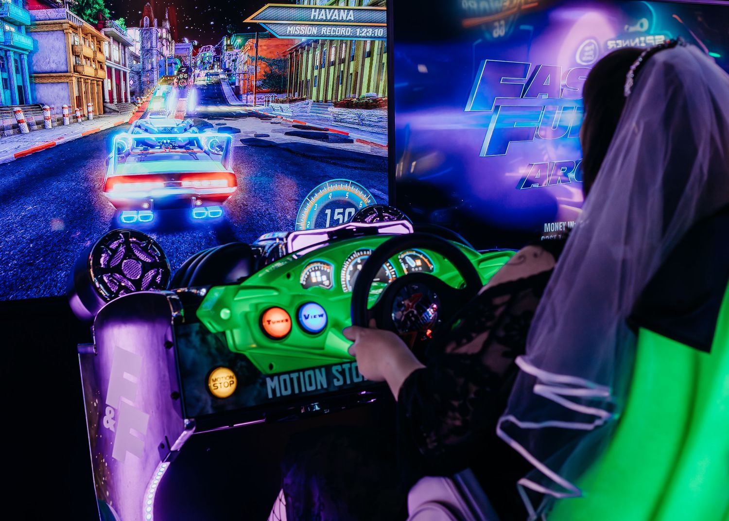 Neon-lit racing arcade game cabinet with bright green car controller and glowing purple lights in a dimly lit room.