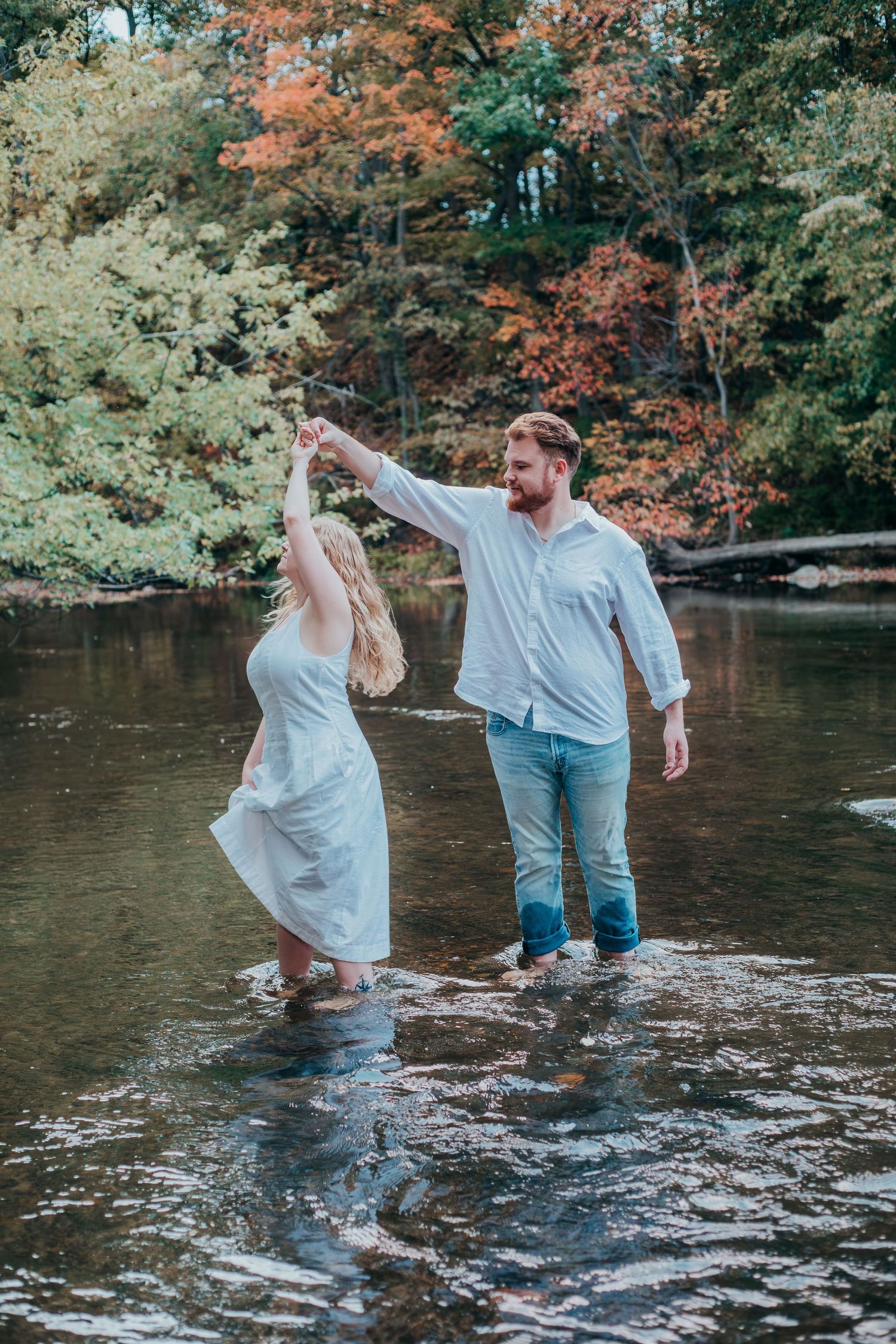 A couple in light clothing plays and splashes together in a scenic river surrounded by autumn foliage.
