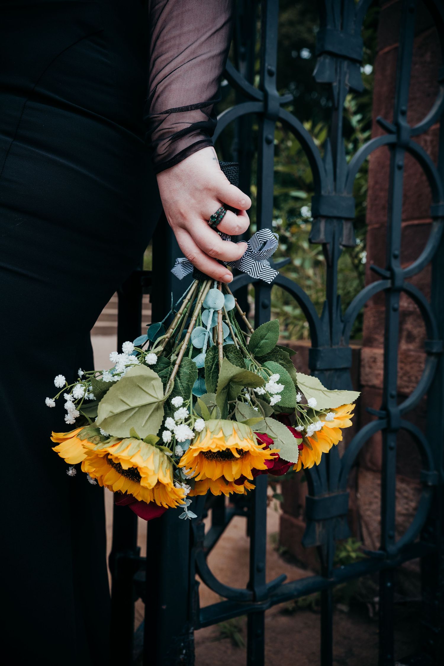 Hand holding fresh sunflowers and baby's breath against dark ornate metal gate in moody lighting.
