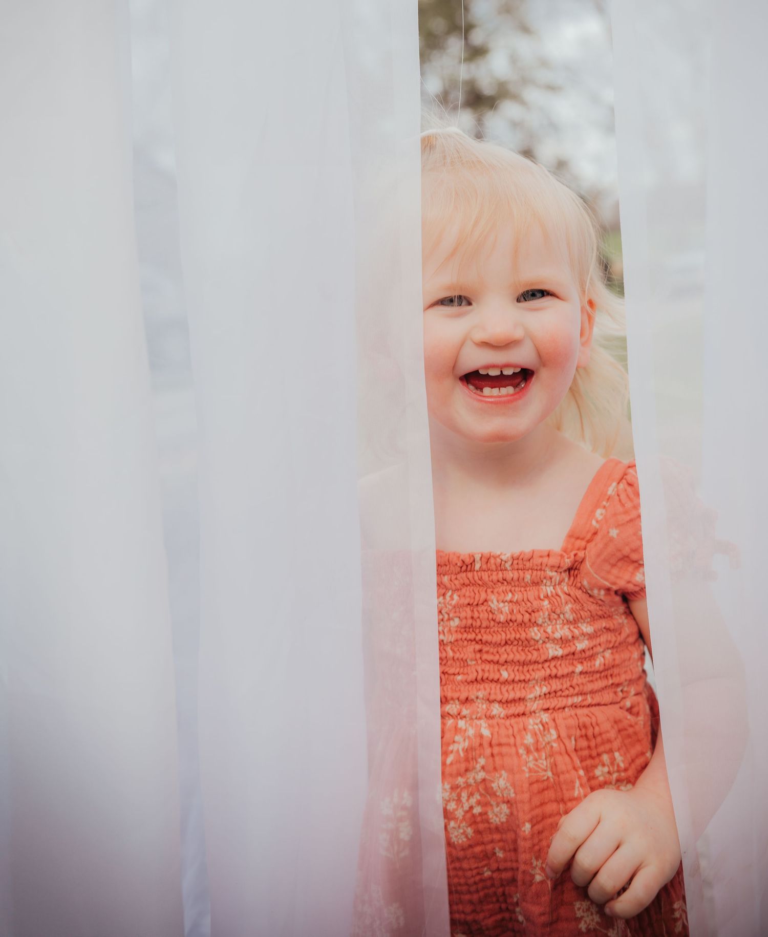 A peek through white curtains showing someone wearing an orange dress with a joyful expression.