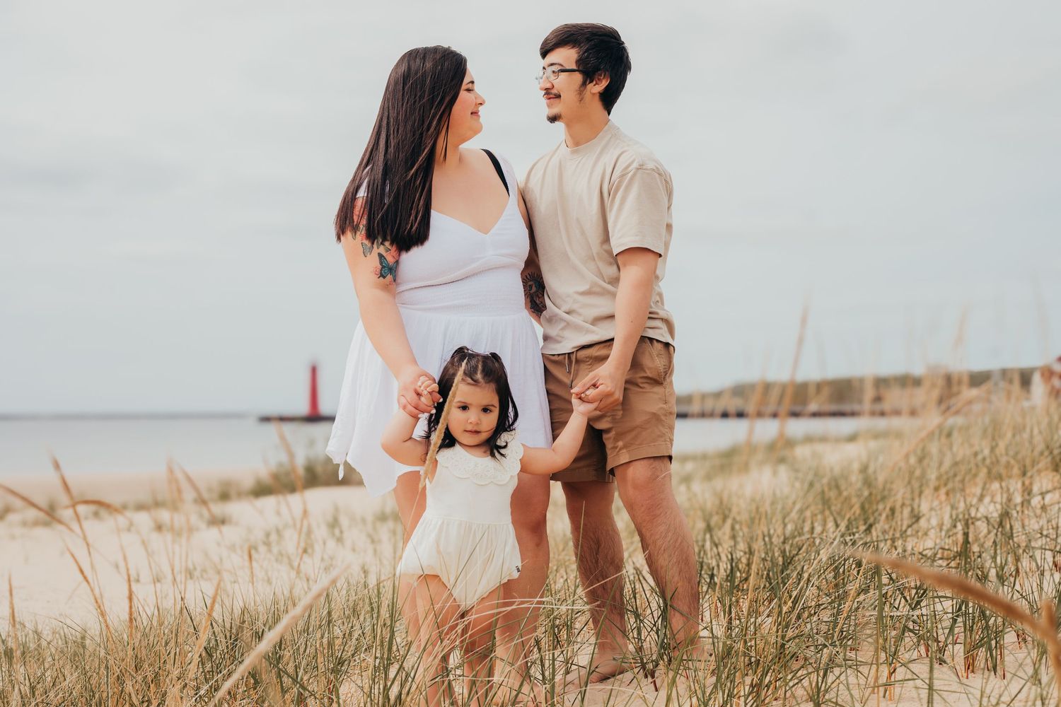 A family stands together on a grassy beach dune with a lighthouse visible in the background on a cloudy day.