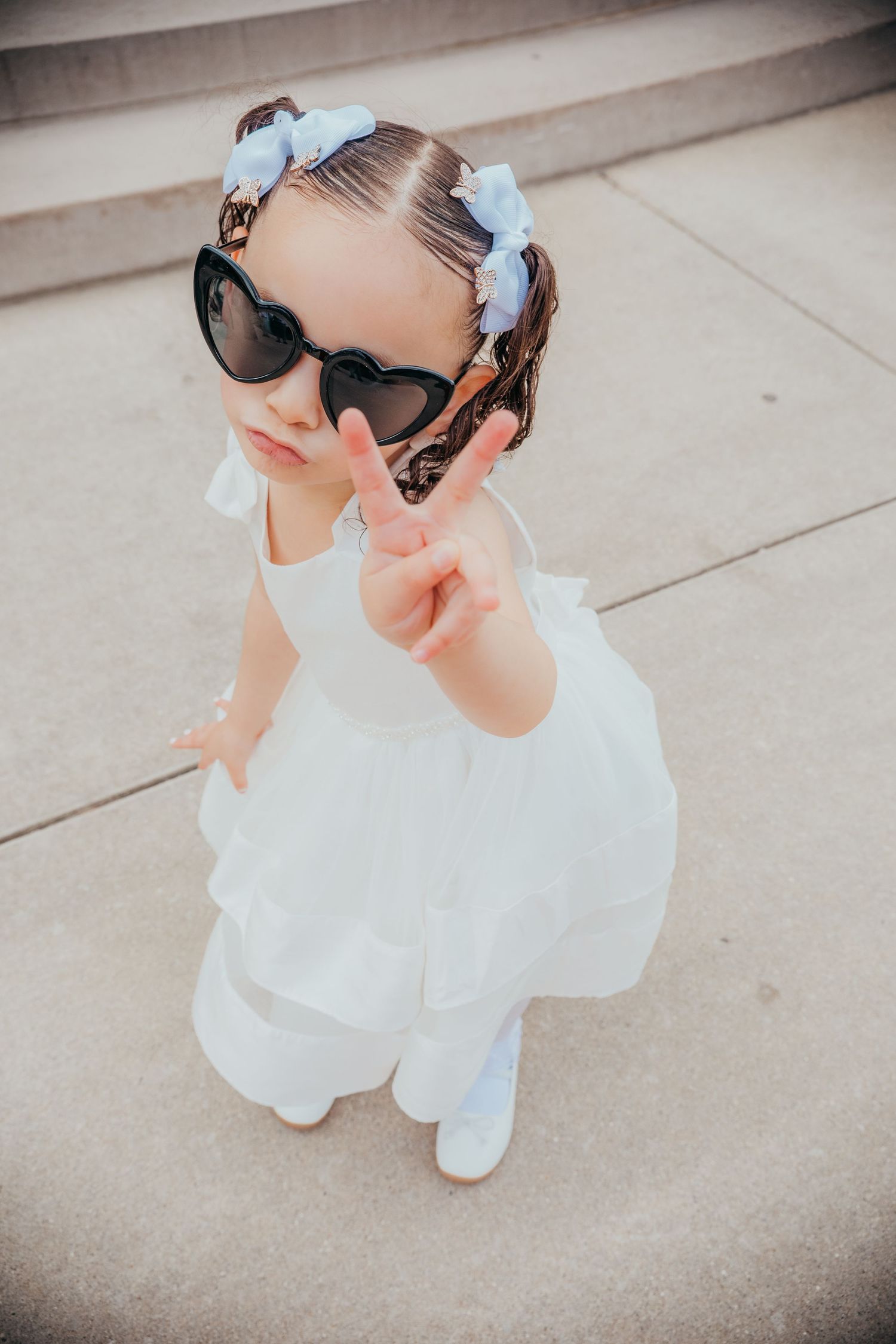 A person in a white dress and sunglasses sits on concrete steps with a light blue bow in their hair.