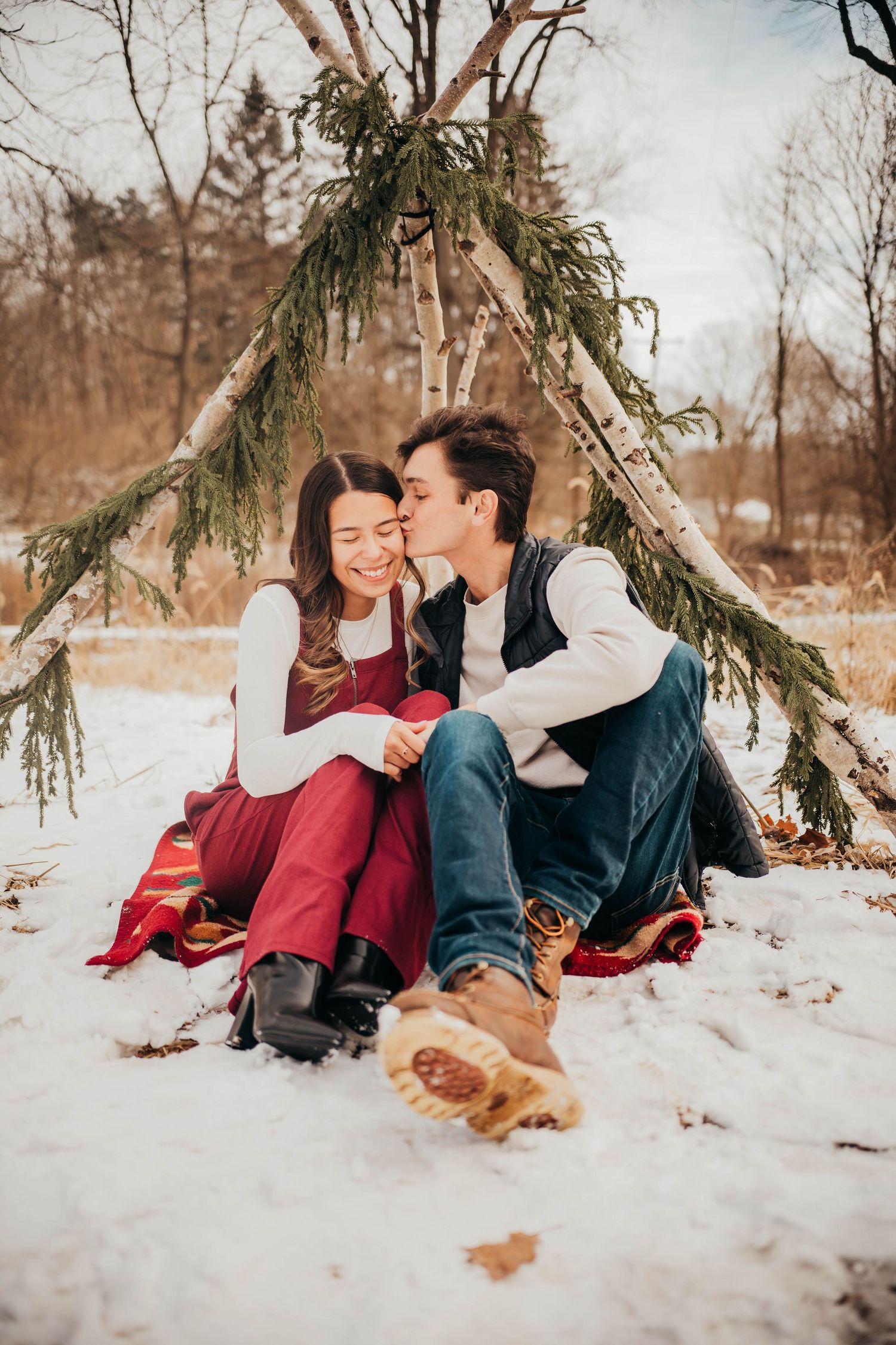 A couple shares an intimate moment while sitting together in the snow under evergreen branches during winter.