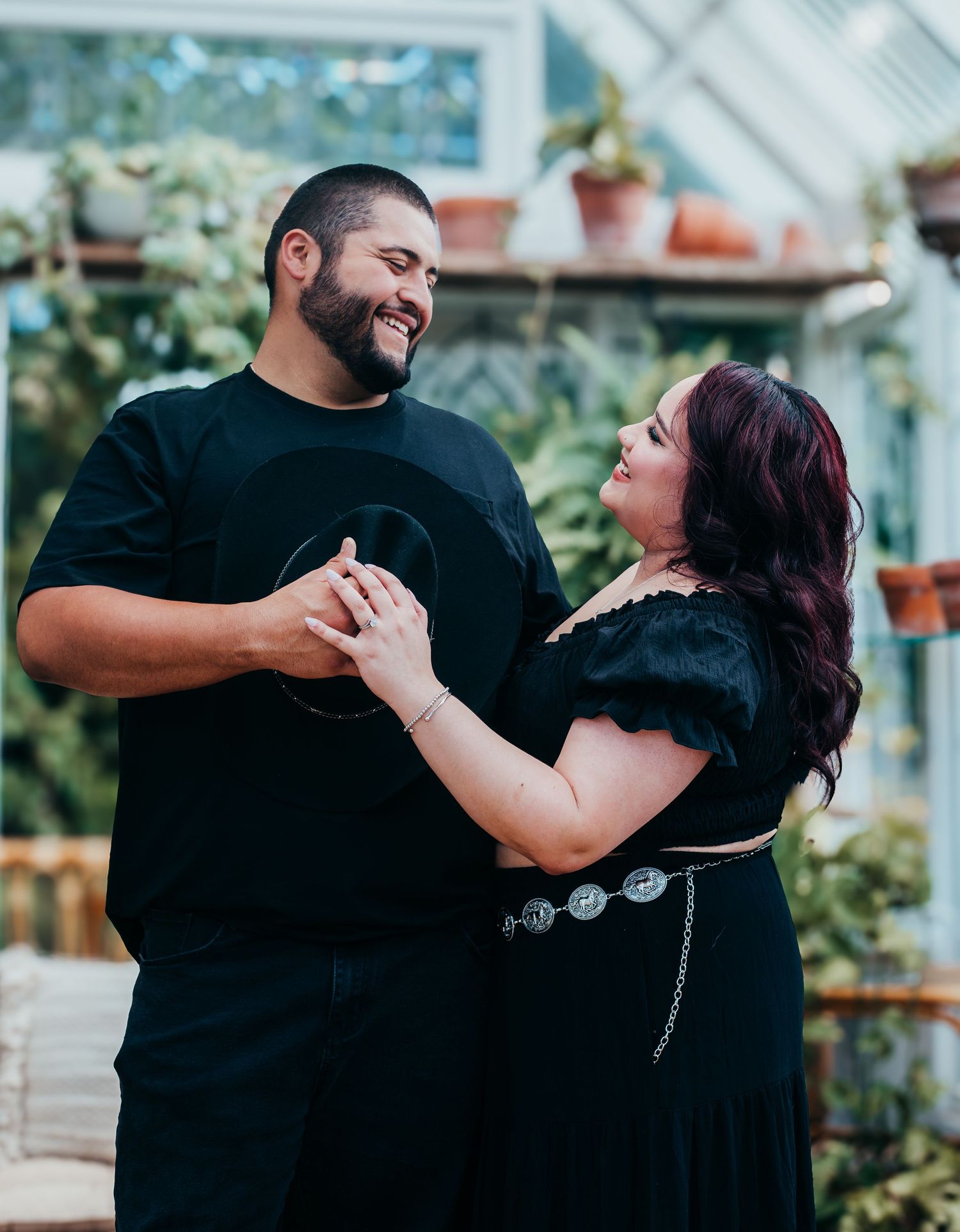 A couple in black clothing embraces and laughs together in a garden setting during a photo session.