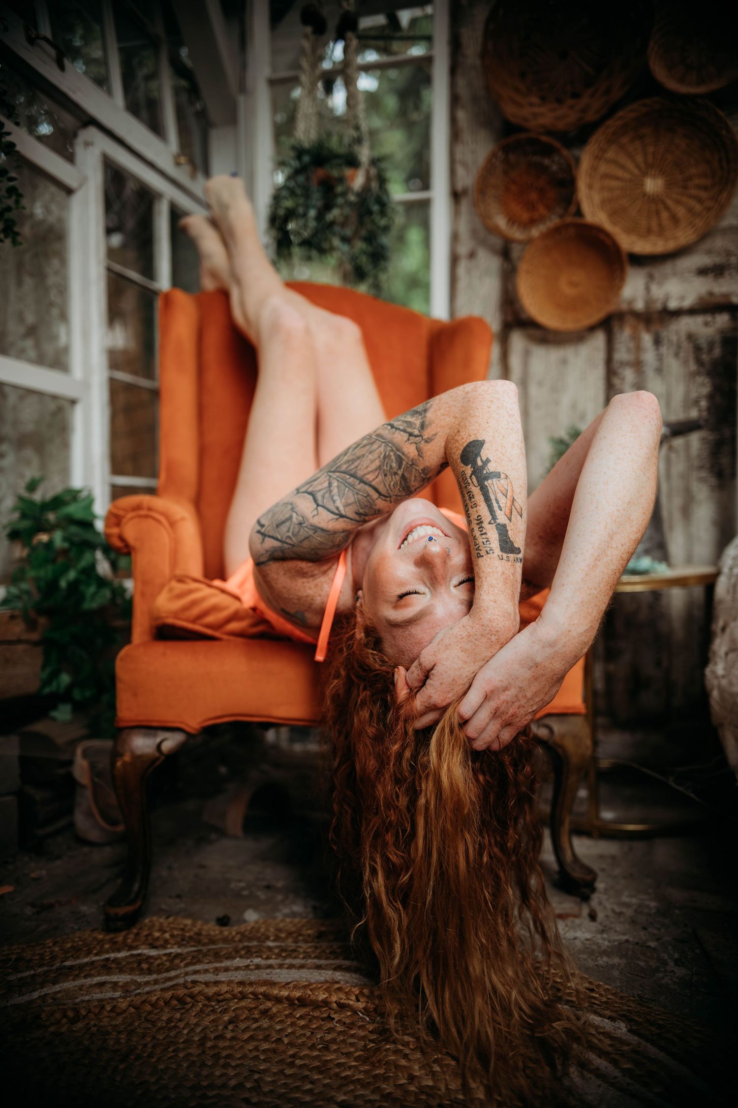 A tattooed figure with long red hair reclines dramatically on an orange chair near decorative woven baskets.