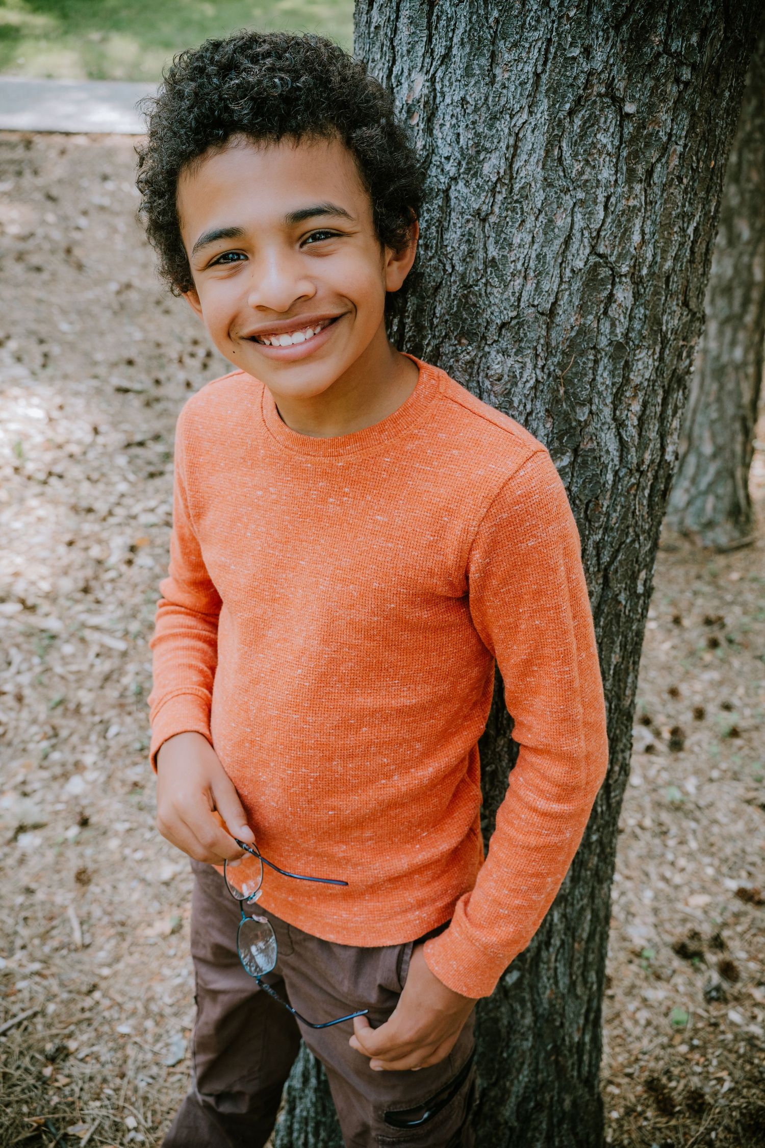 A child in an orange sweater and brown pants plays near a tree trunk in a series of candid outdoor photos.