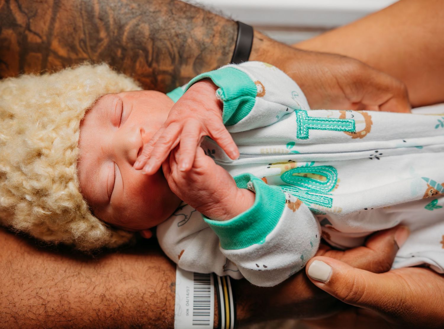 Infant wearing a white and teal outfit being gently held by caring hands.