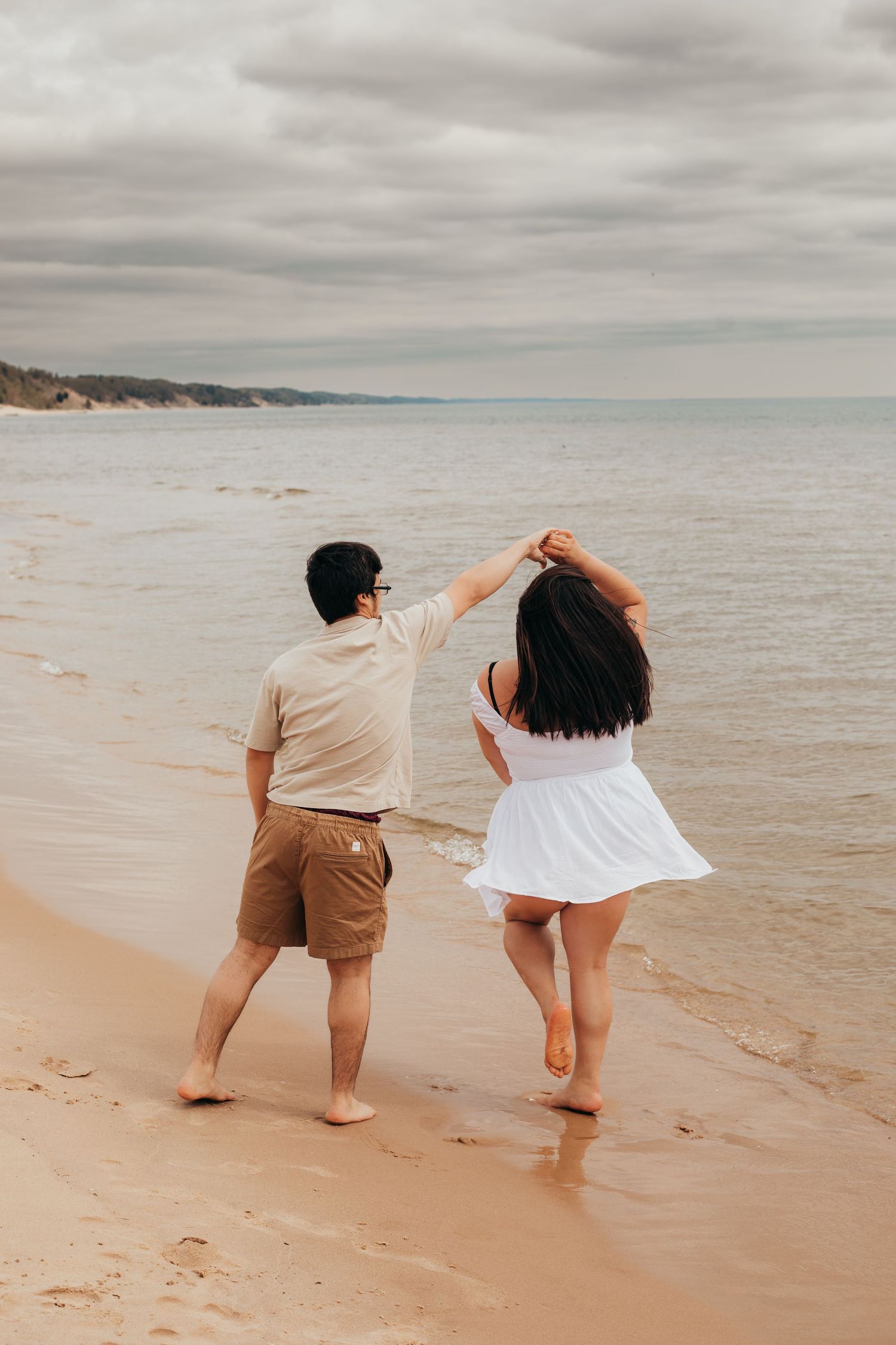 A couple in casual attire playfully dips their toes in the ocean waves along a serene sandy beach on a cloudy day.