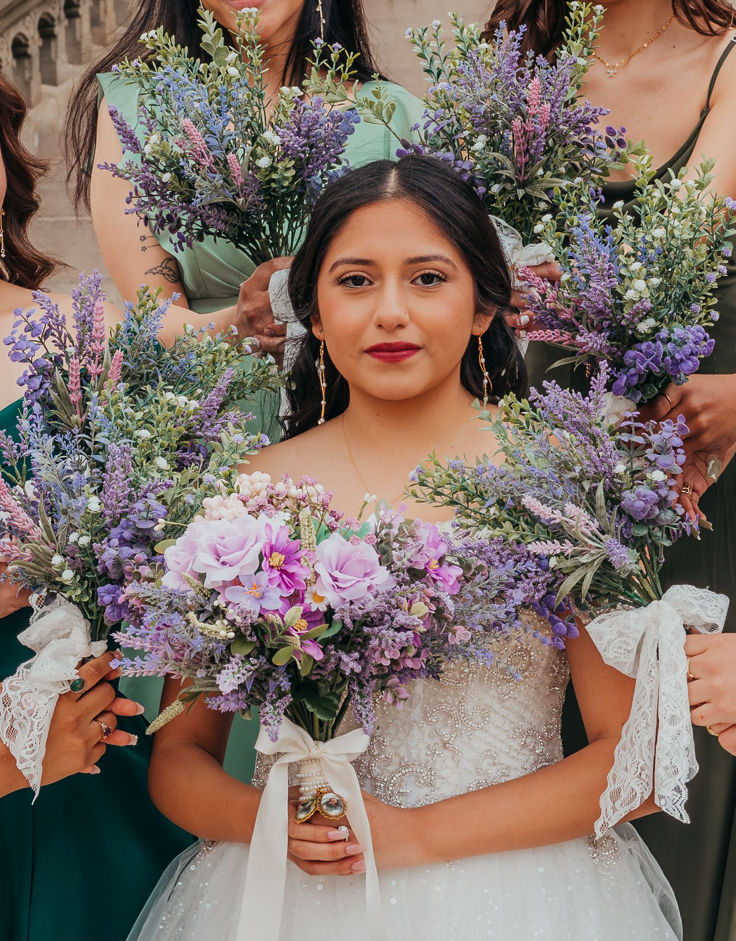 Wedding bouquet features lavender and purple wildflowers with soft pink roses against a white lace dress.