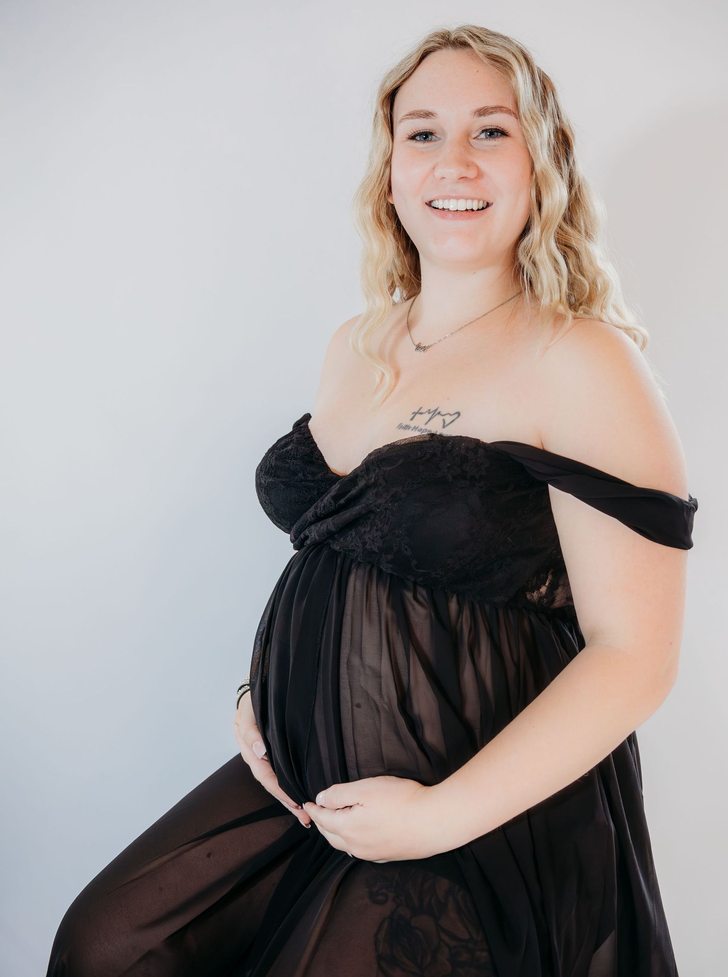 A pregnant woman in a black off-shoulder dress poses against a light background for a maternity photo.