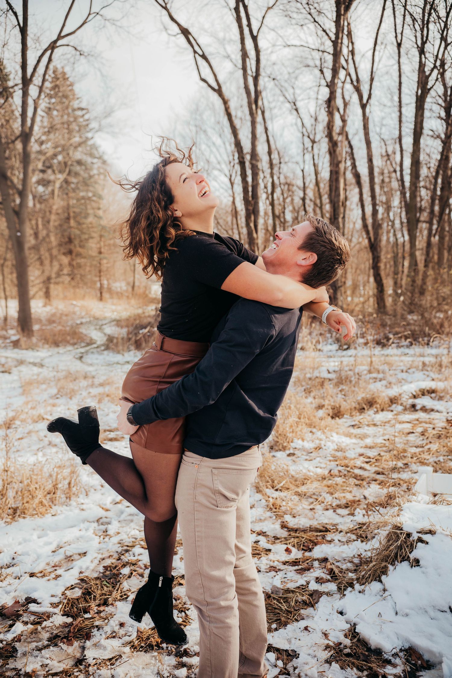 A couple embraces joyfully in a snowy winter forest setting with bare trees in the background.