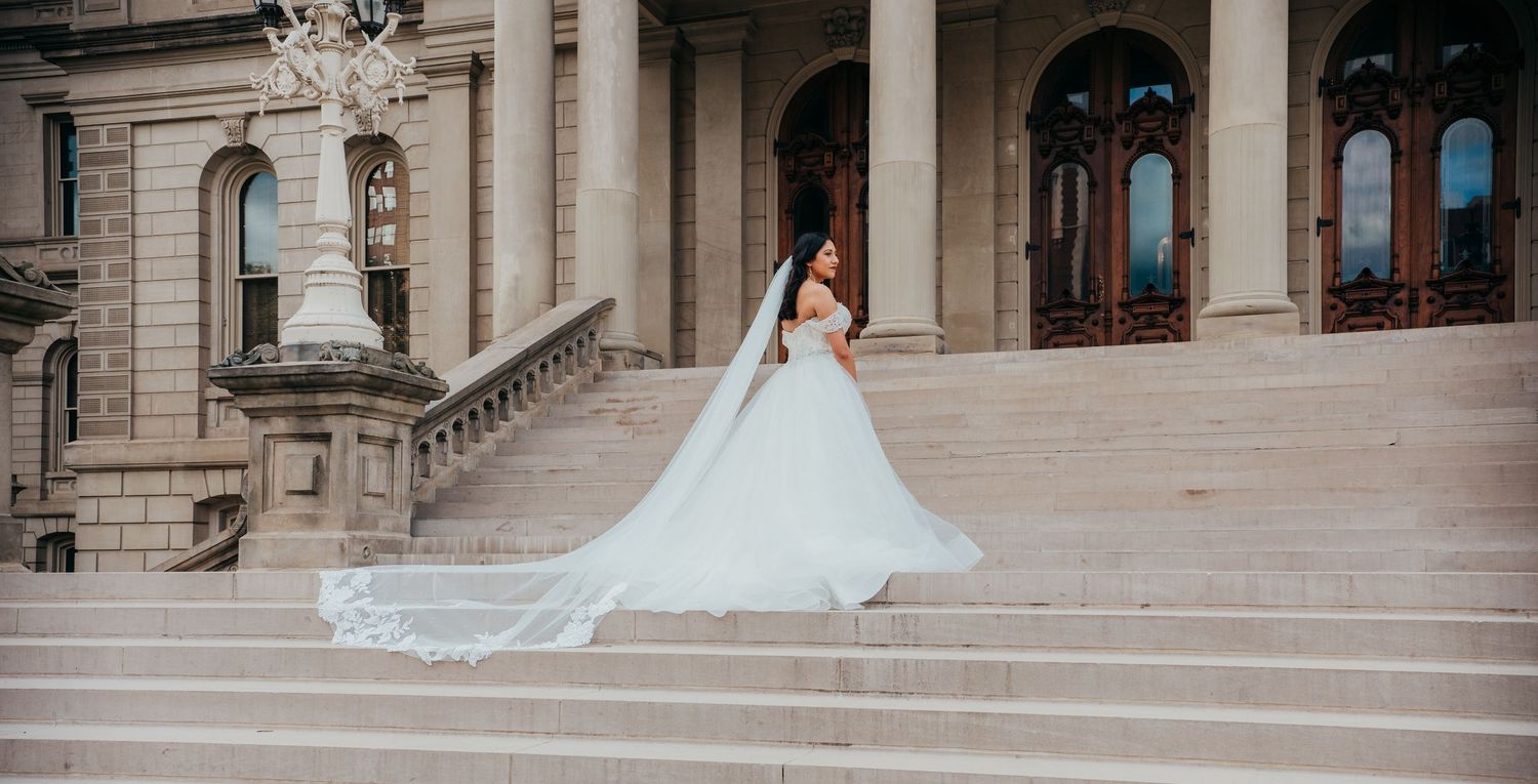 A bride in a flowing white wedding gown poses gracefully on grand marble steps of an elegant historic building.