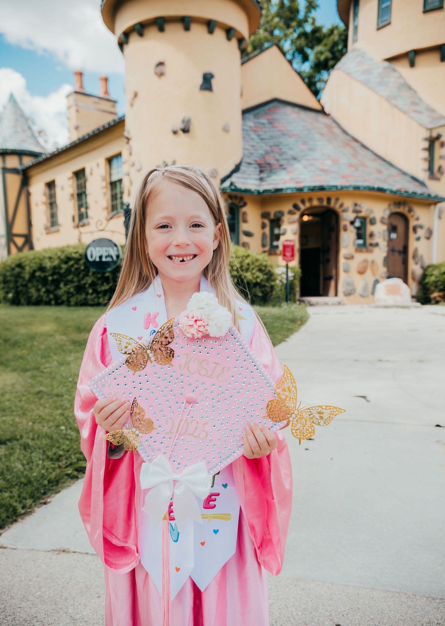 A child in a pink princess dress holds a decorative heart with gold butterflies outside a fairy tale-style castle building.